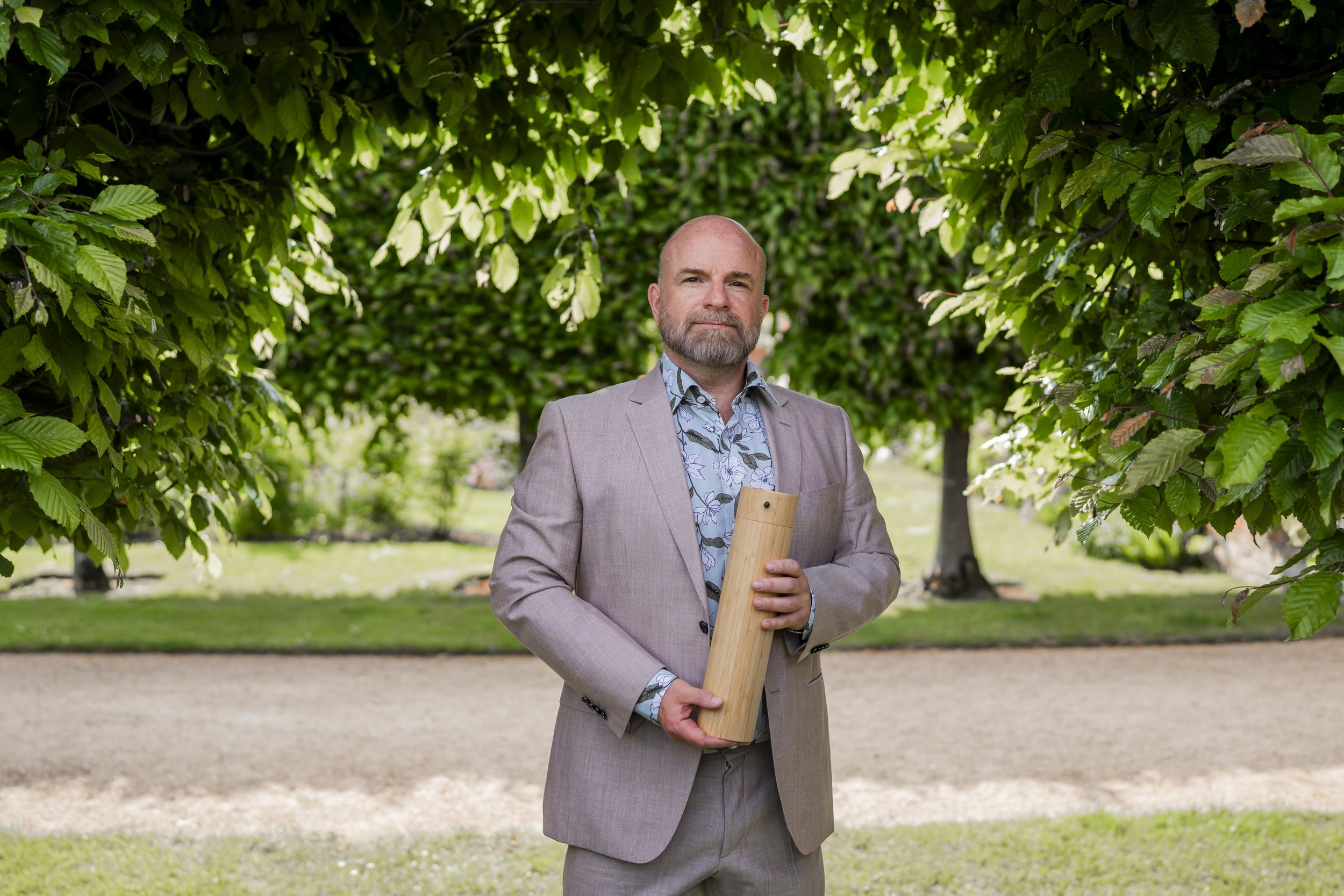 A man with a beard wearing a suit and a floral short holding a bamboo urn standing in front of trees