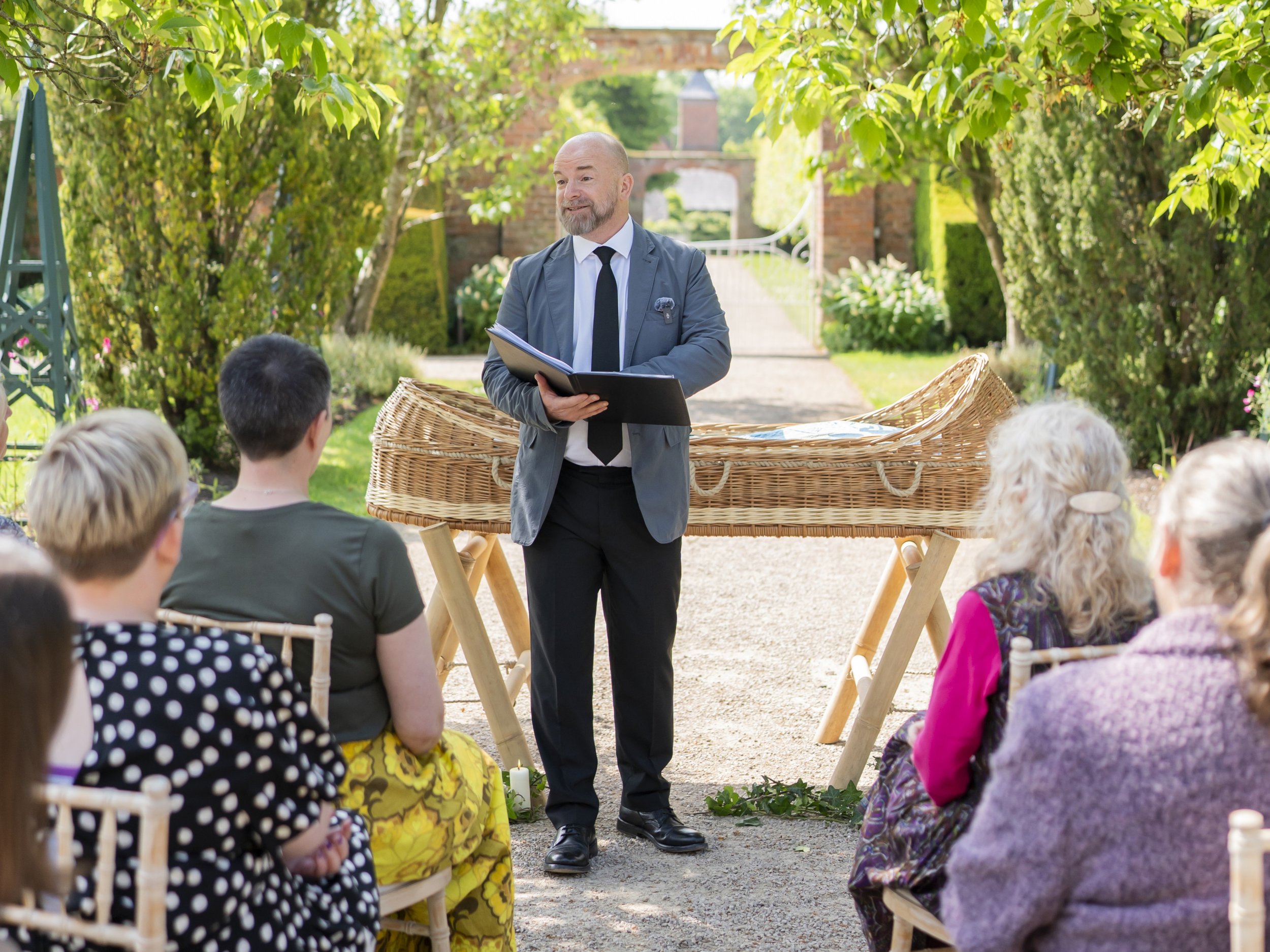 A man with a beard wearing a suit and a black tie holding a book and standing in front of a coffin in a garden speaking to people seated on chairs