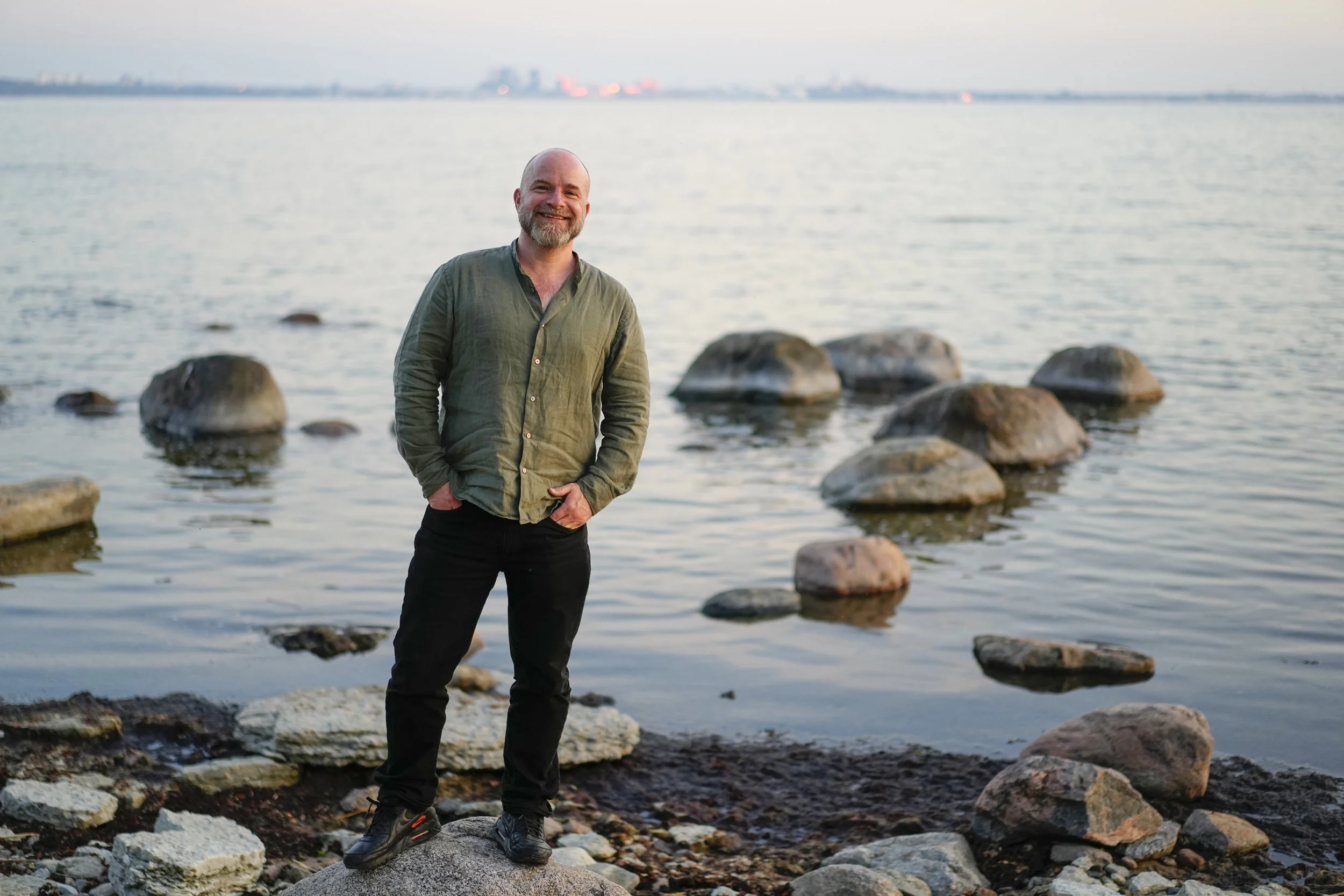 A smiling man wearing a green shirt standing on rocks at the edge of the sea