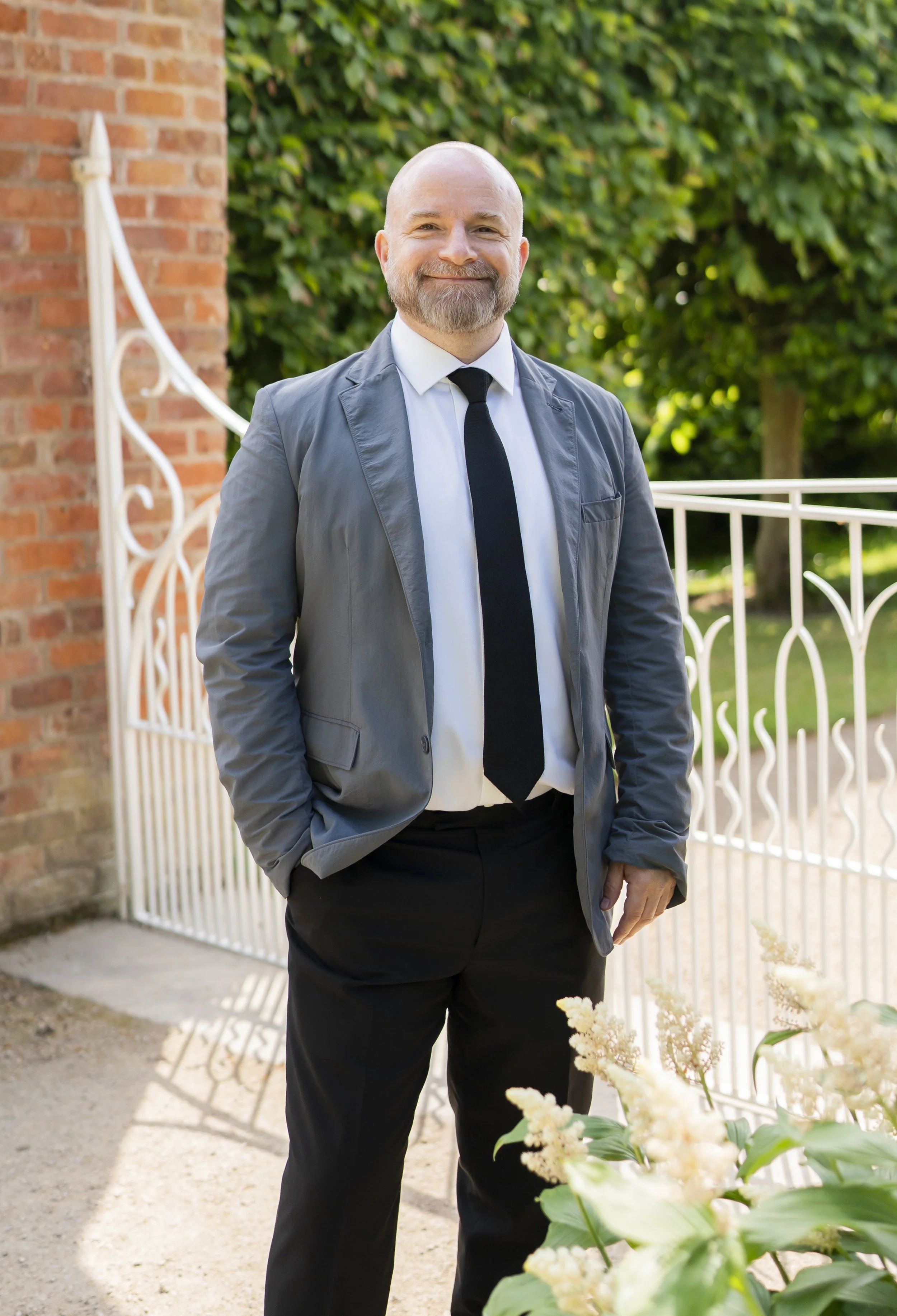 A smiling man with a beard wearing a suit and a black tie with one hand in a trouser pocket standing in front of a gate