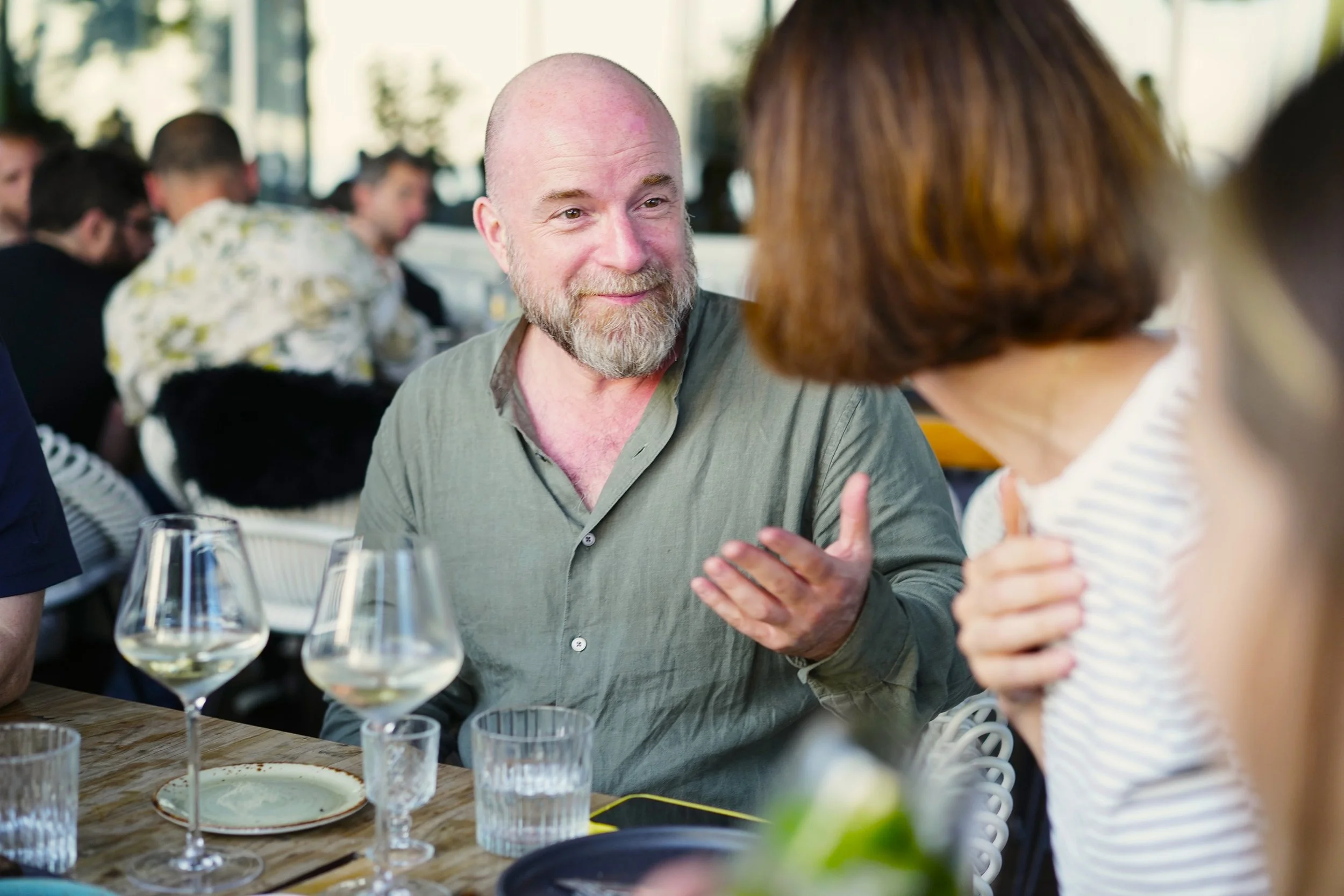 A man with a beard wearing a green shirt sitting at a table with an outstretched hand speaking to another person