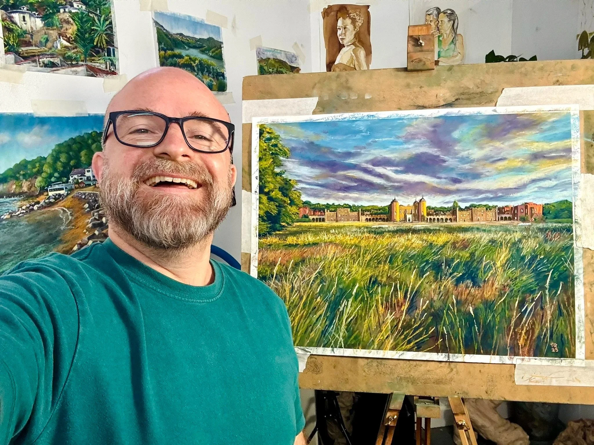 A smiling man with a beard and glasses wearing a green shirt standing in front of a painting of a landscape