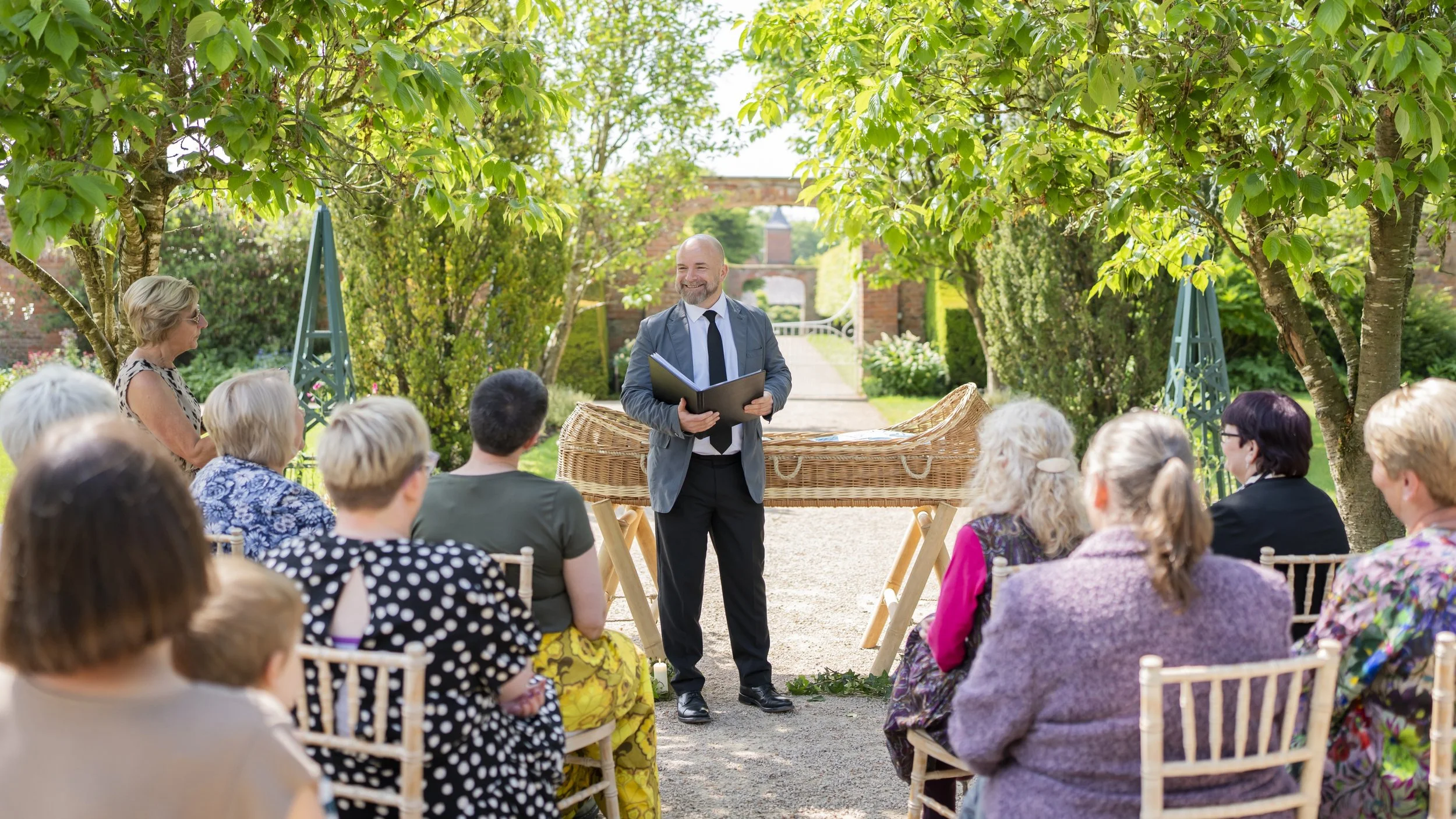 A man wearing a suit and black tie holding a book standing in front of a coffin at a funeral in a garden with people sitting on chairs watching