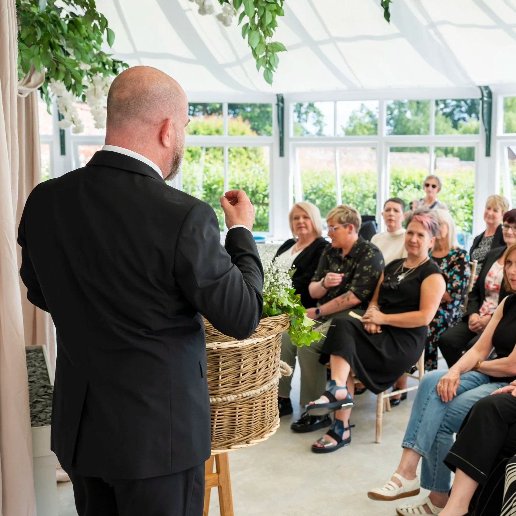 The back of a man wearing a black suit speaking to people seated on chairs at a funeral