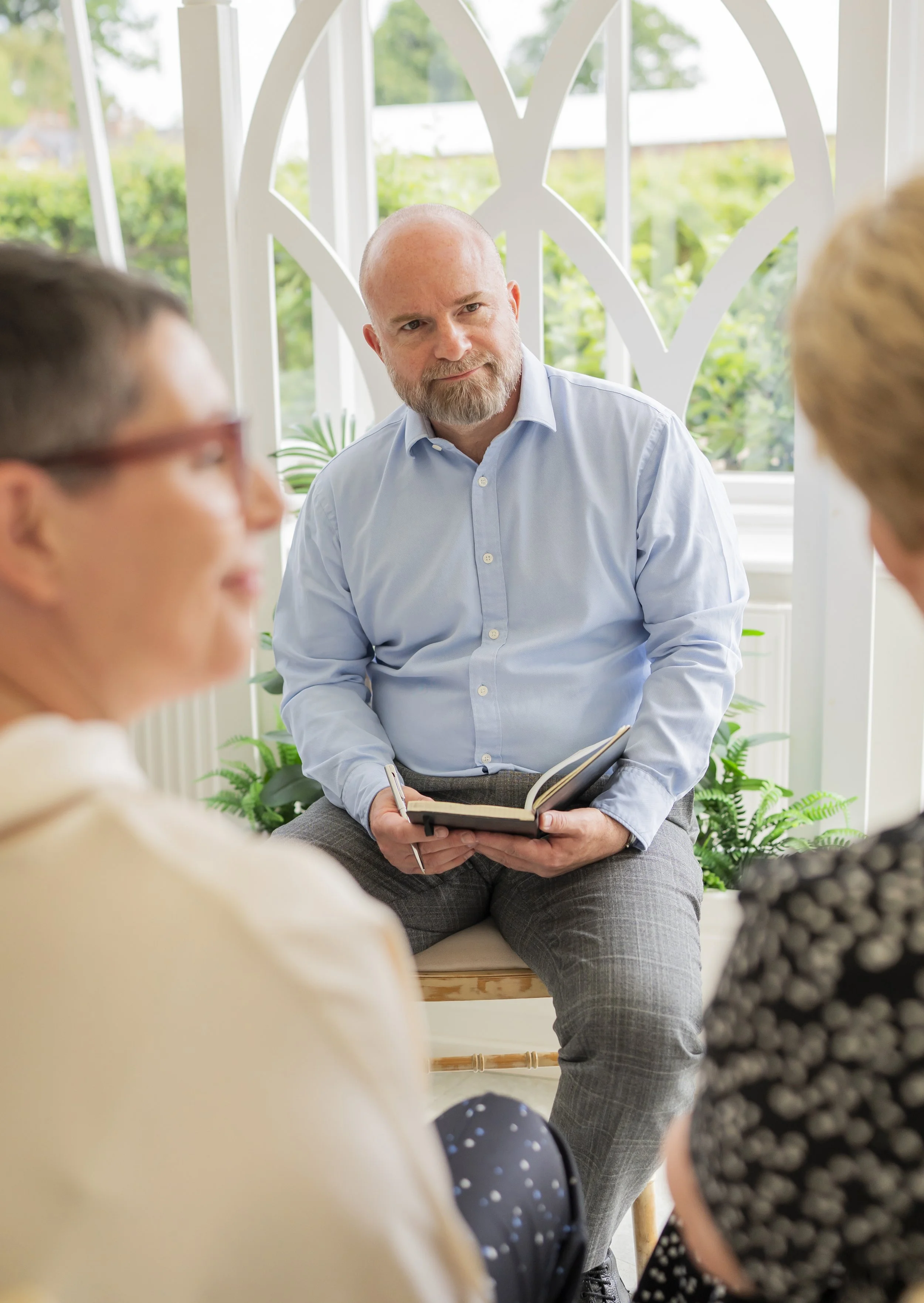 A seated man with a beard wearing a blue shirt holding a notebook and pen listening to another person