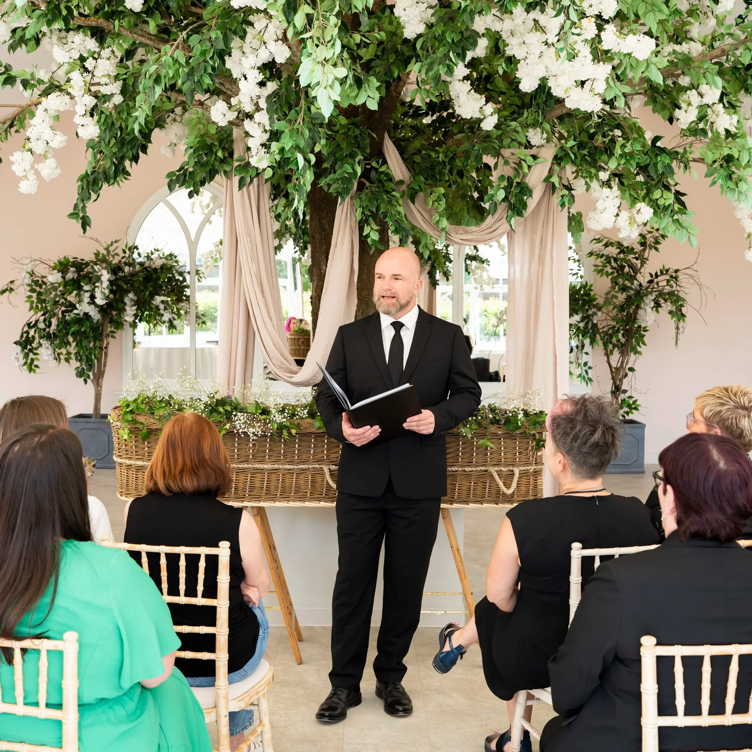 A man with beard wearing a black suit and black tie holding a book and standing in front of a wicker coffin at a funeral ceremony