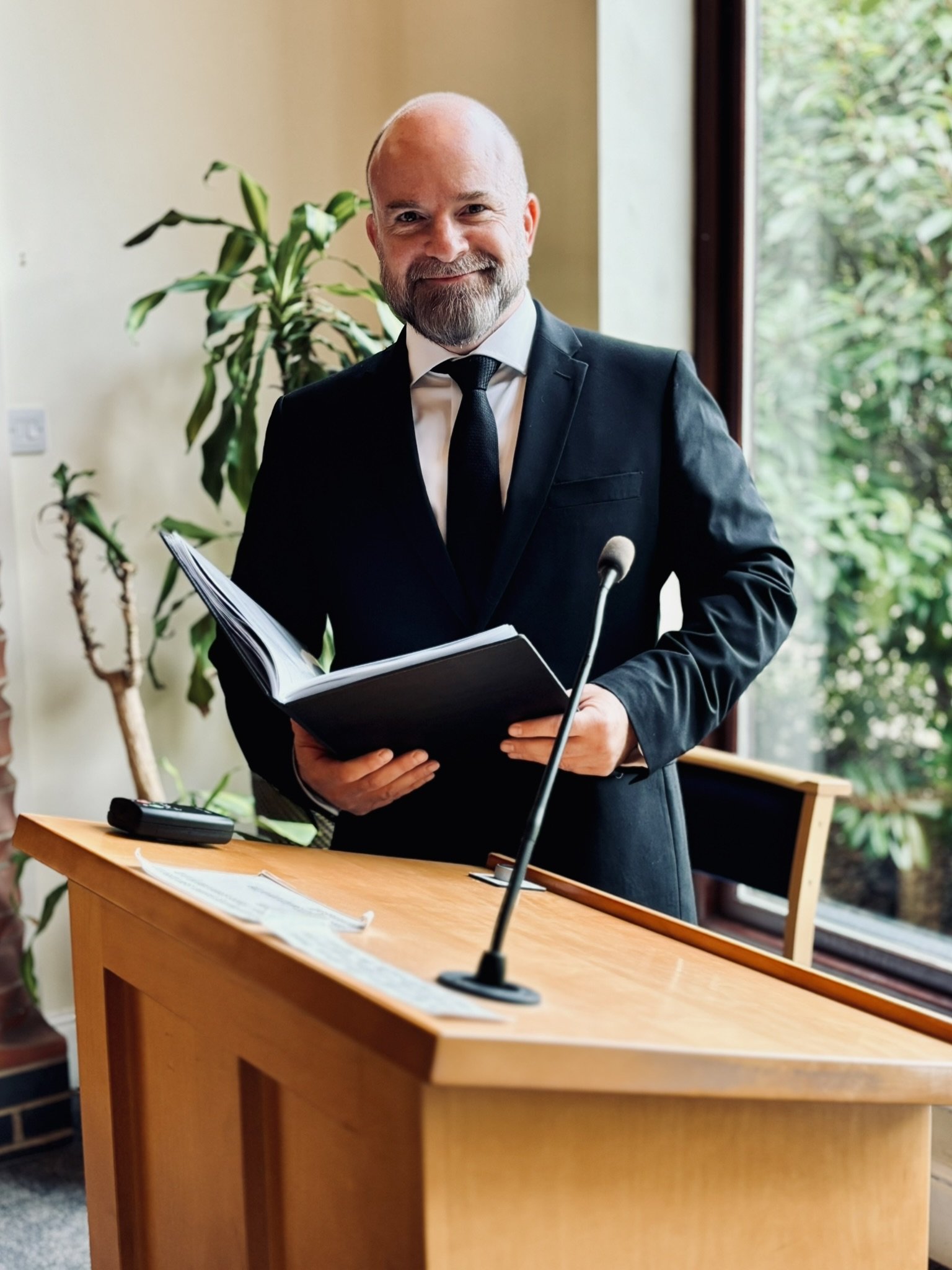 A man with beard wearing a black suit and black tie standing at a podium holding a book