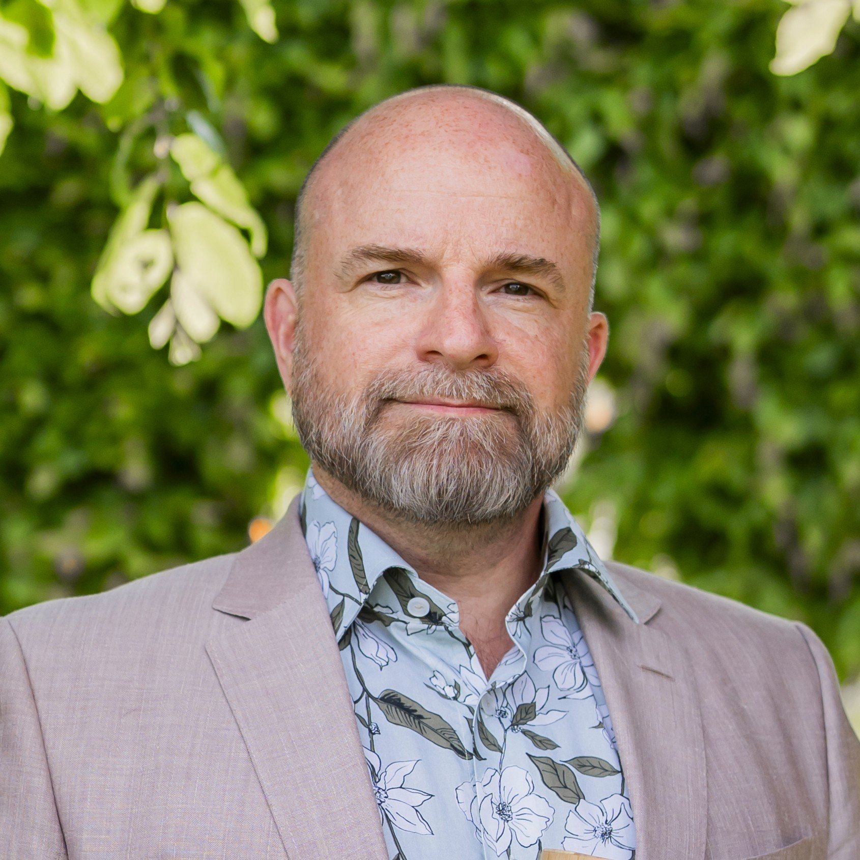 A man with beard wearing a beige jacket and floral shirt standing in front of green foliage