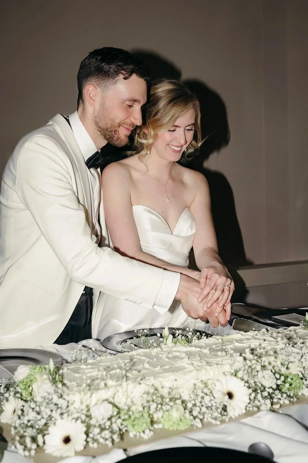 A newlywed bride and groom in wedding attire cutting a wedding cake together, smiling, with white floral decorations on the cake.