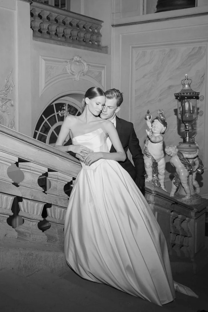 A black and white editorial wedding photograph of a bride and groom dressed elegantly, standing closely on a grand staircase in a historic building with ornate decoration in Residenzschloss Ludwigsburg.