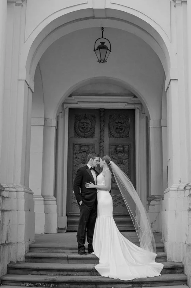 Black and white editorial wedding photo of a bride and groom kissing on the steps of an ornate building with classical architecture and decorative columns in Residenzschloss Ludwigsburg.