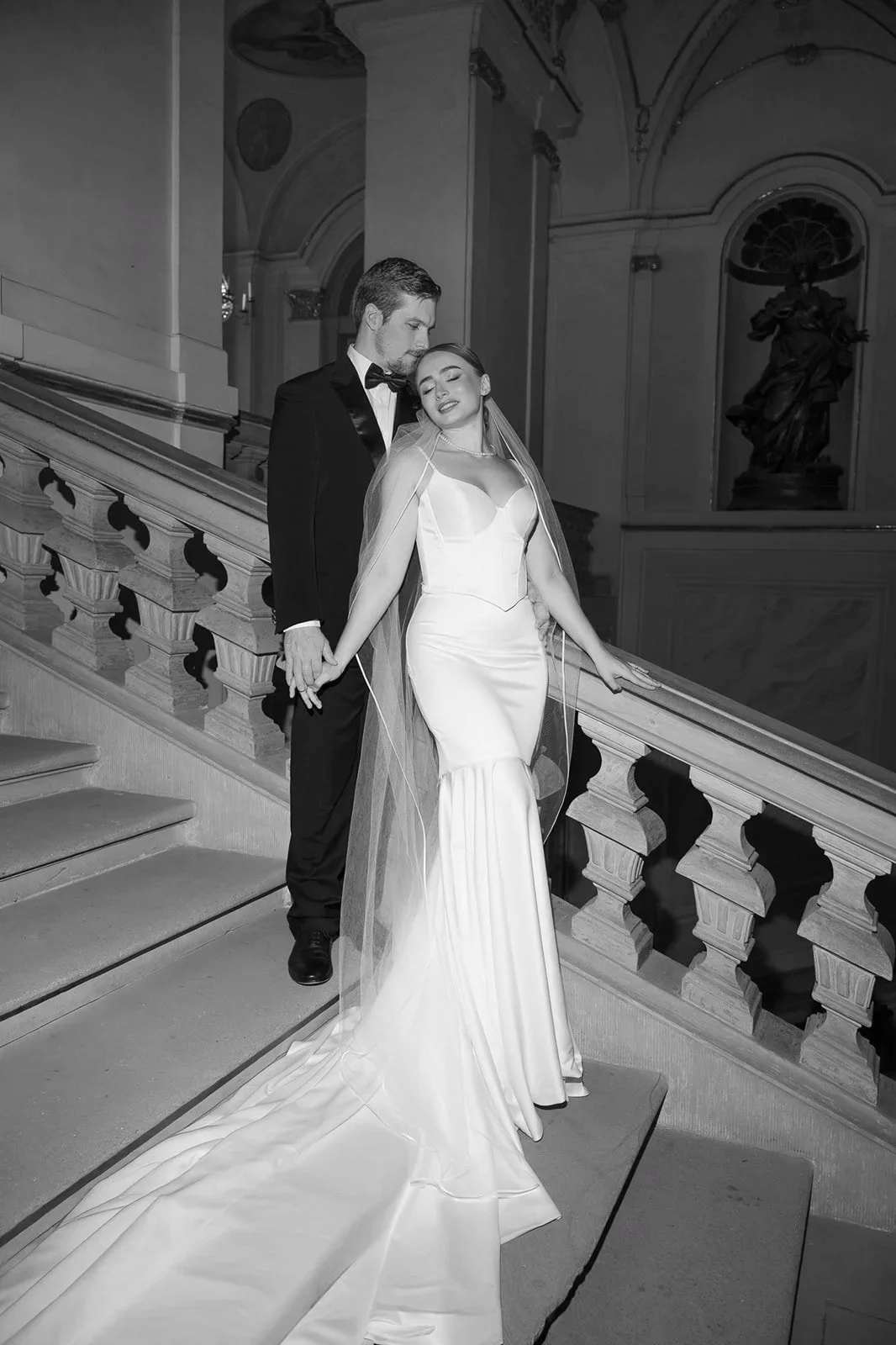 A bride and groom holding hands on a staircase during their wedding, with the bride in a white gown and the groom in a tuxedo. The setting appears to be a grand, elegant building with statues and ornate interior design in Residenzschloss Ludwigsburg.