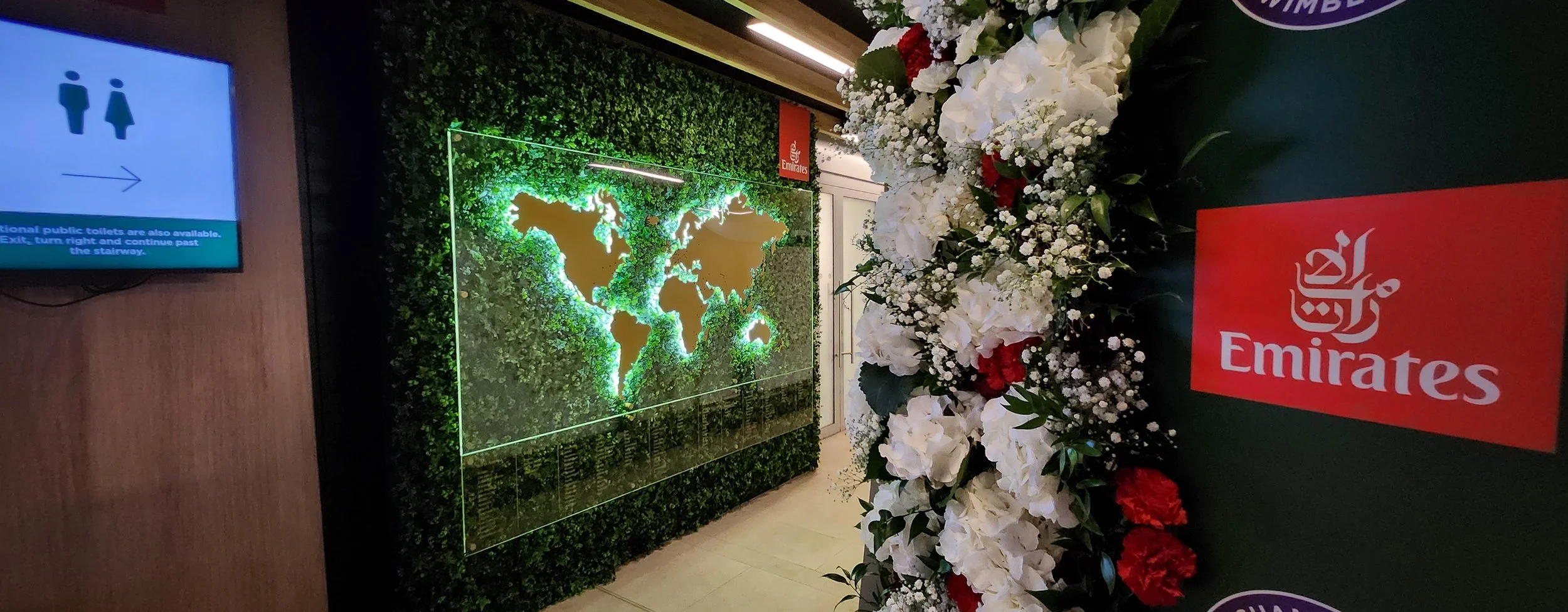 A wall decorated with a lush green plant arrangement featuring a world map outline illuminated with green lights. In front of the wall, there is a bouquet of white and red flowers and a red Emirates logo on a black surface.