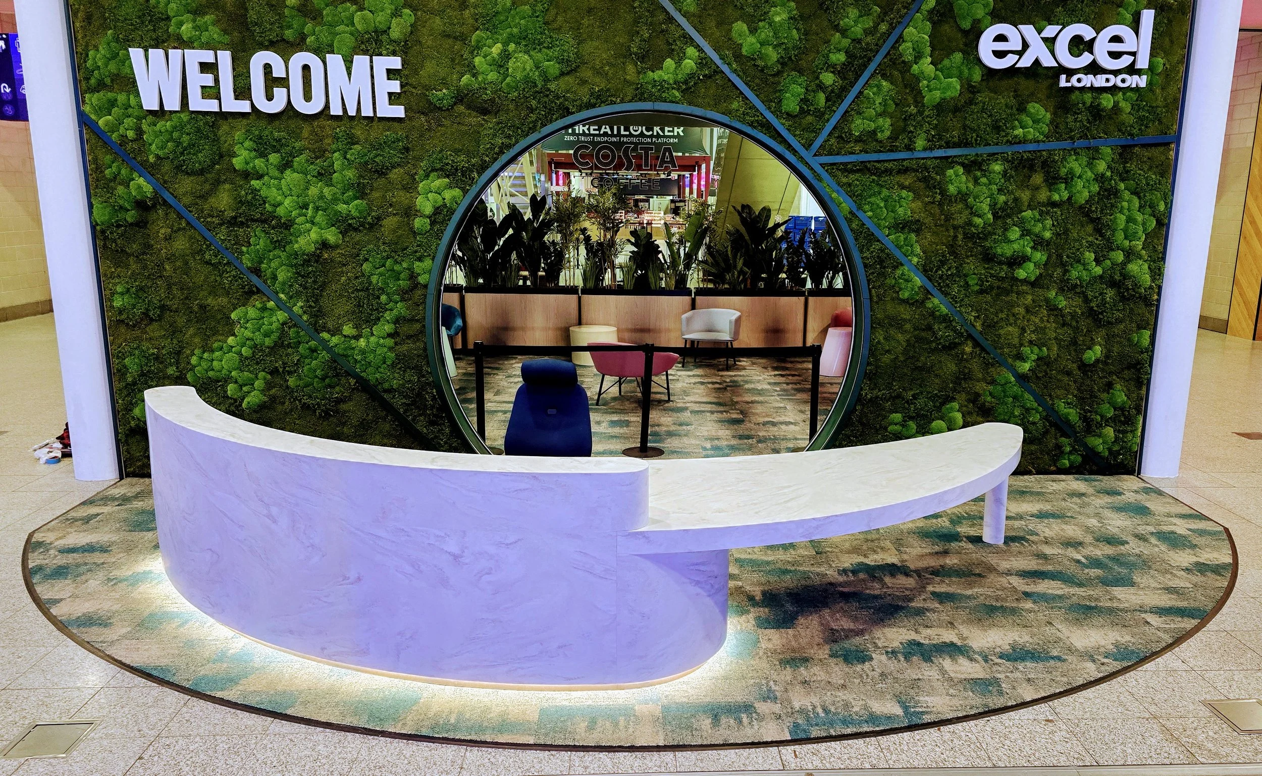 Modern reception desk in front of a green moss wall with the words 'WELCOME' and 'excel London' displayed. Behind the desk, there is a circular mirror showing a seating area with chairs and plants inside a shopping mall.