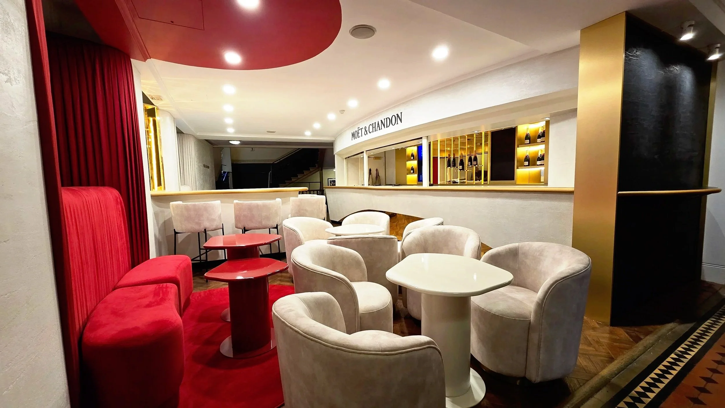 Interior of a luxurious lounge area with cream-colored armchairs, a red velvet bench, and a red table, with a Moët & Chandon bar in the background surrounded by gold accents.