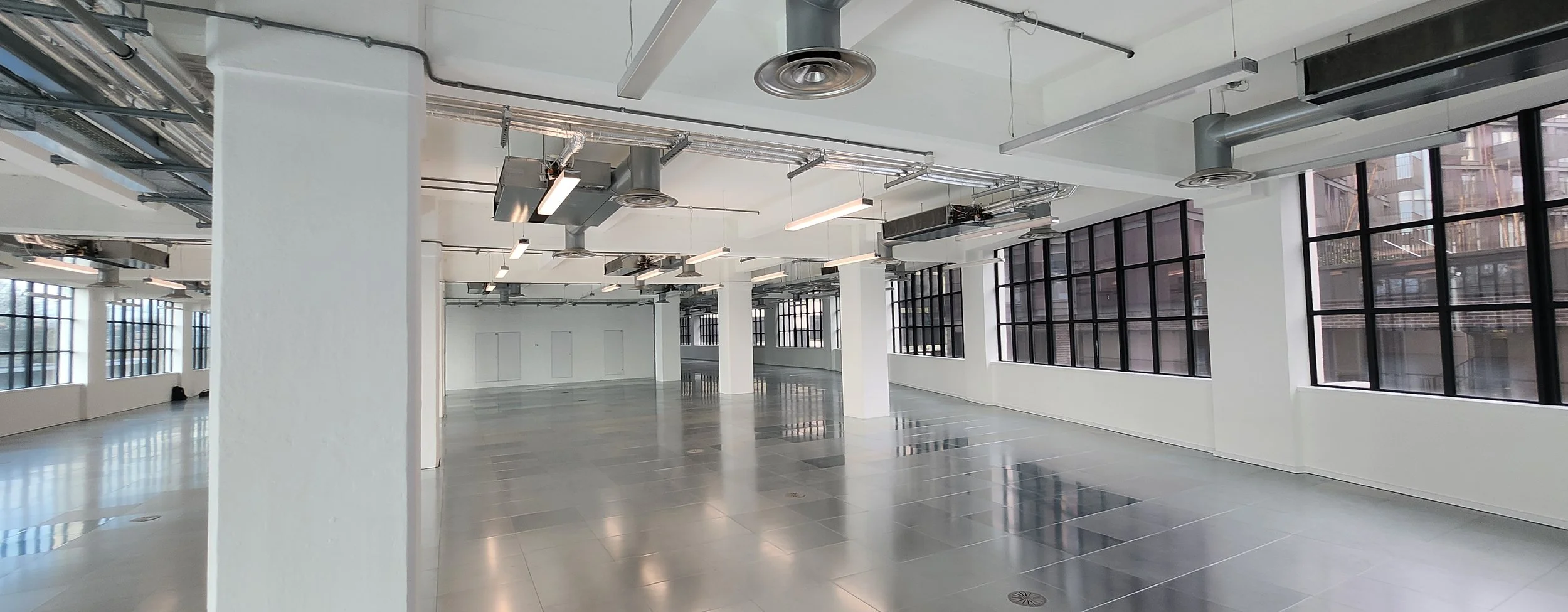 Empty office space with large windows, exposed ductwork, and modern lighting fixtures.