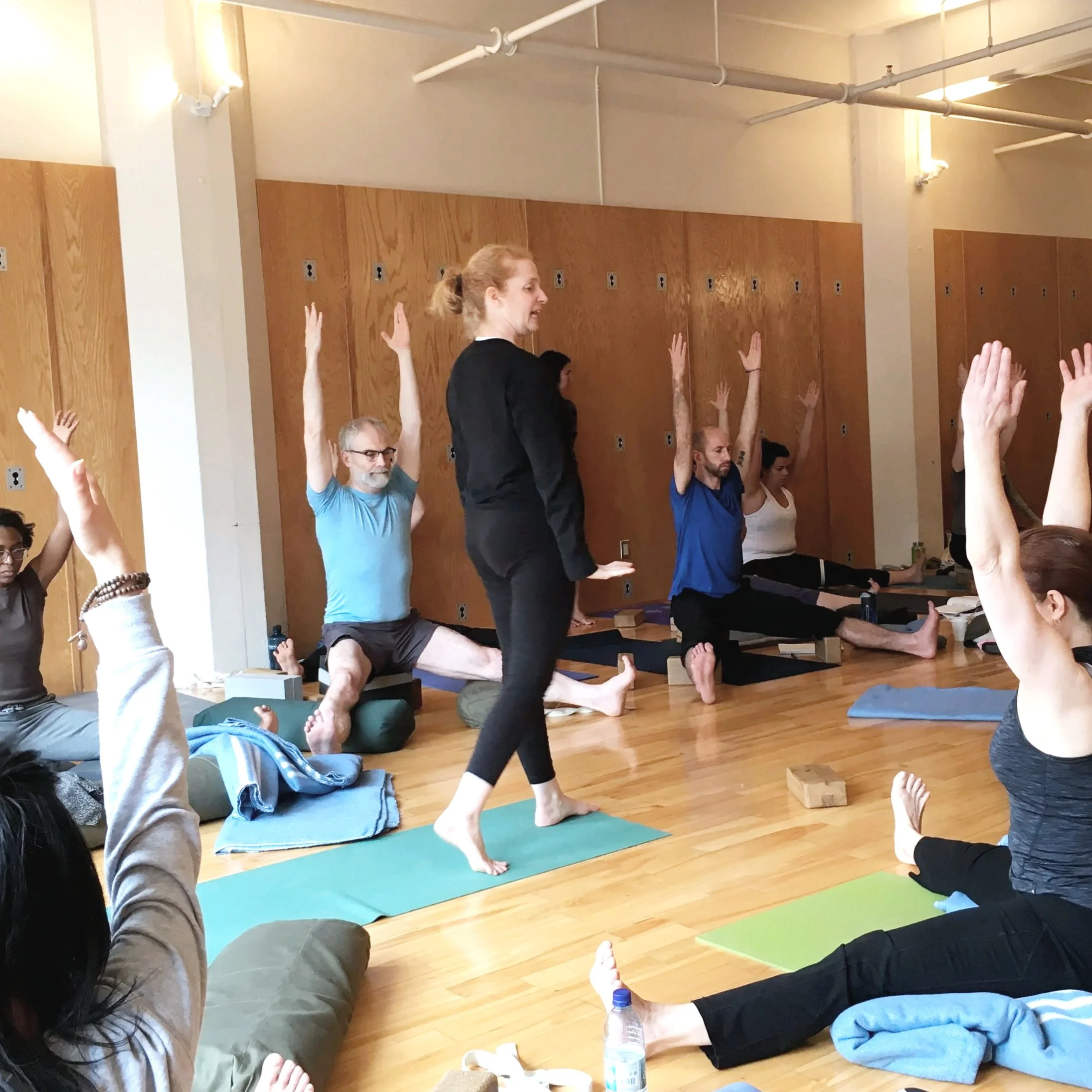 Participants in a yoga class are sitting on yoga mats with their arms raised. An instructor is standing and demonstrating a pose in a wooden-floored studio with wooden wall panels.