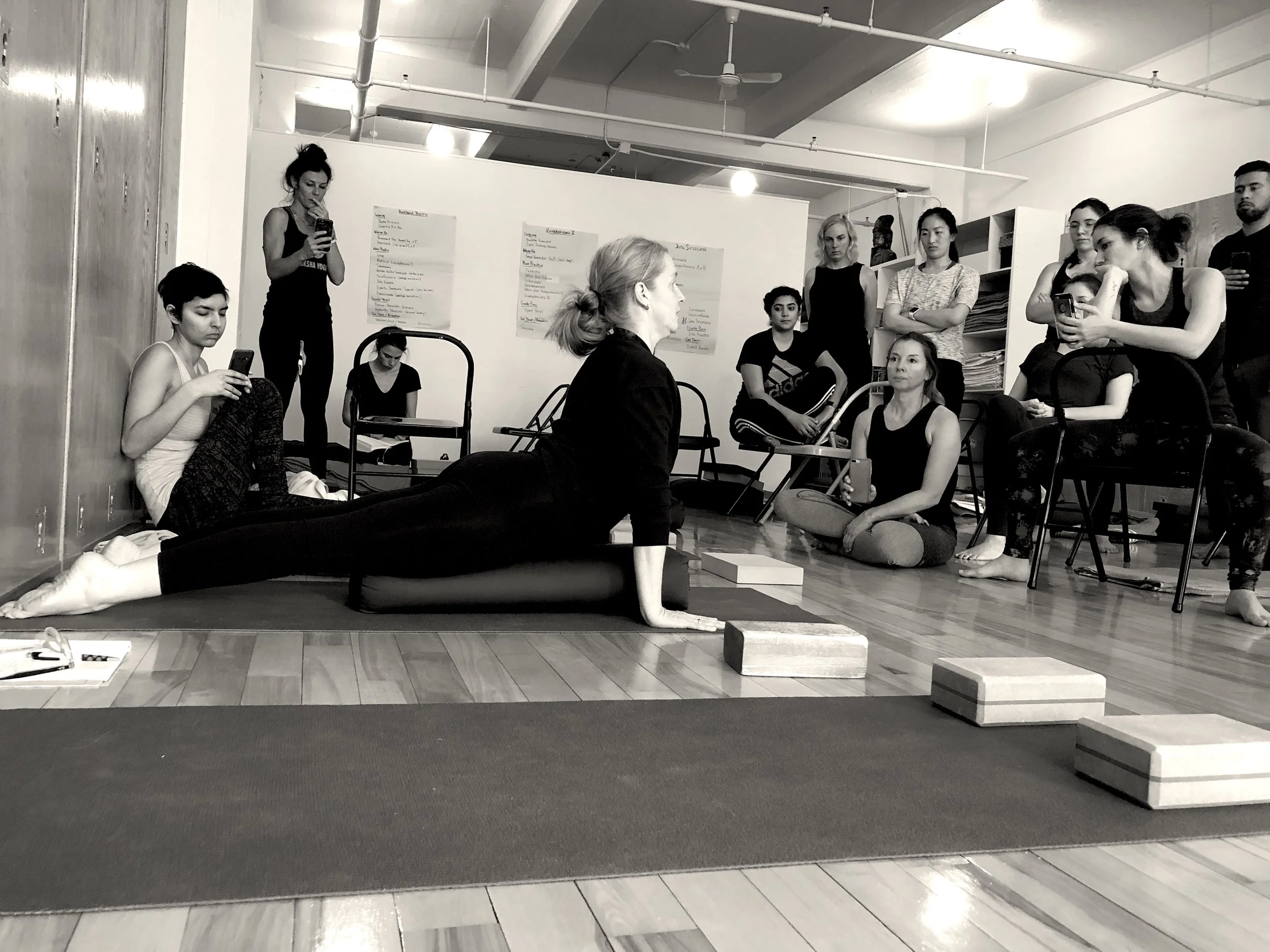 A yoga instructor demonstrates a pose on a yoga mat in front of a group of women, with some seated and others standing, inside a studio with wooden floors and white walls.