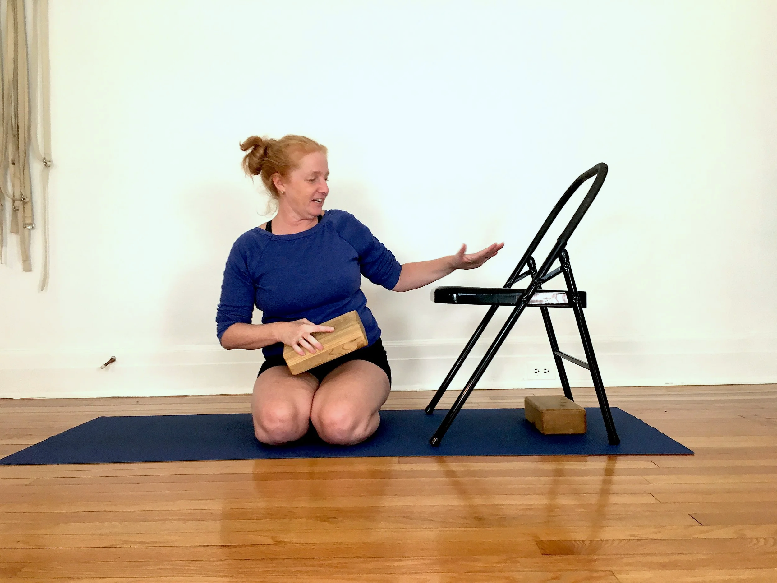 A woman kneeling on a yoga mat with wooden blocks, performing an exercise with a chair on a hardwood floor.