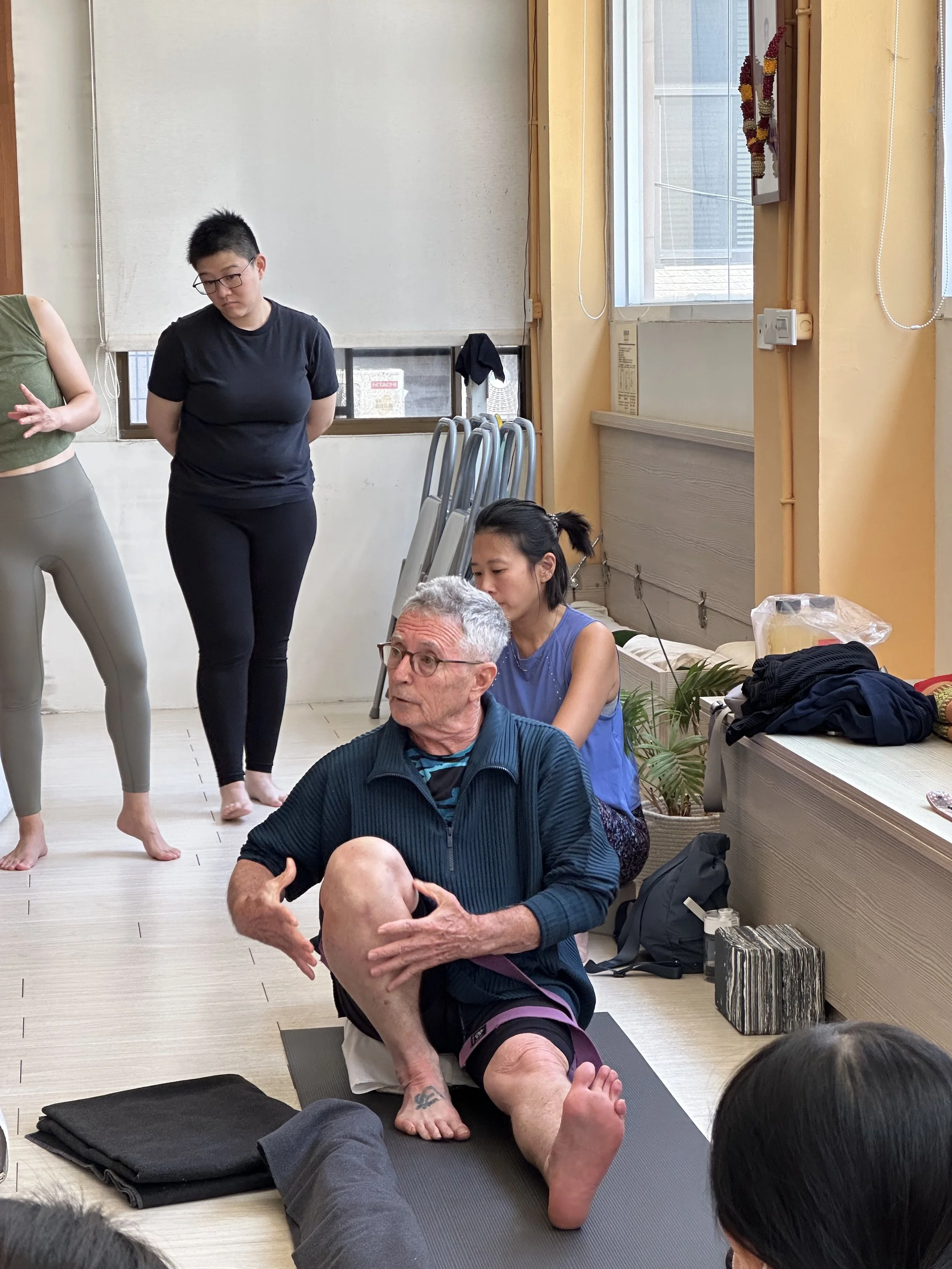An elderly man with gray hair and glasses practices a seated yoga pose while three women observe and assist him in a yoga class. The setting appears to be a yoga studio with mats, chairs, and windows bringing natural light into the room.