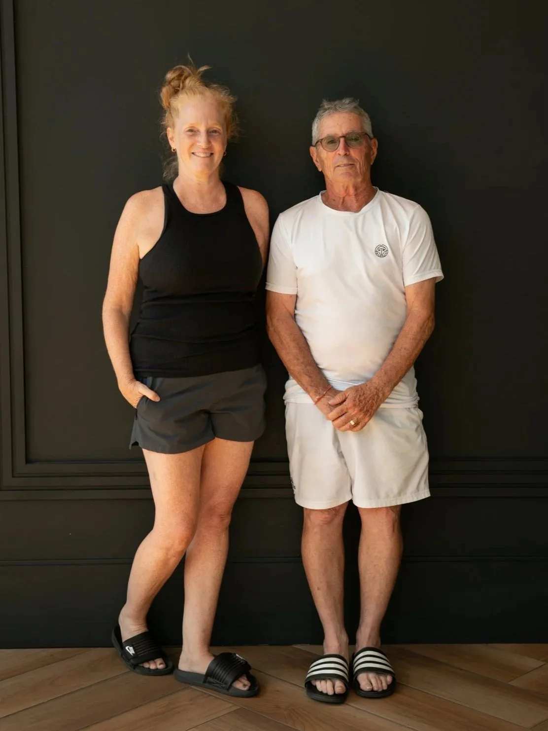 A casually dressed woman and an elderly man stand against a dark wall. The woman wears a black sleeveless top, gray shorts, and black slide sandals. The man is dressed in a white t-shirt, white shorts, and black-and-white striped slide sandals.
