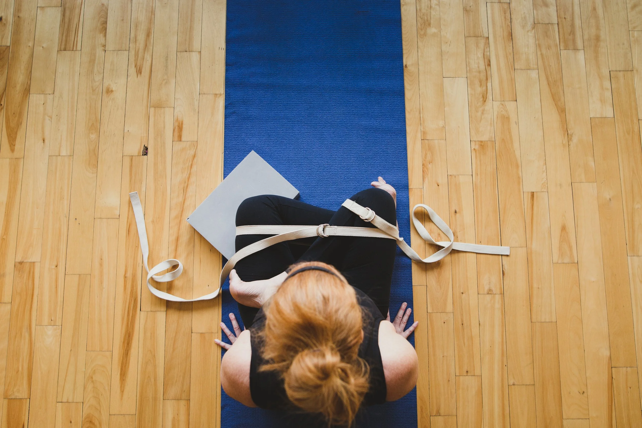 A person with red hair practicing yoga on a blue mat in a wooden-floored room, sitting cross-legged with a gray book or notebook beside them.