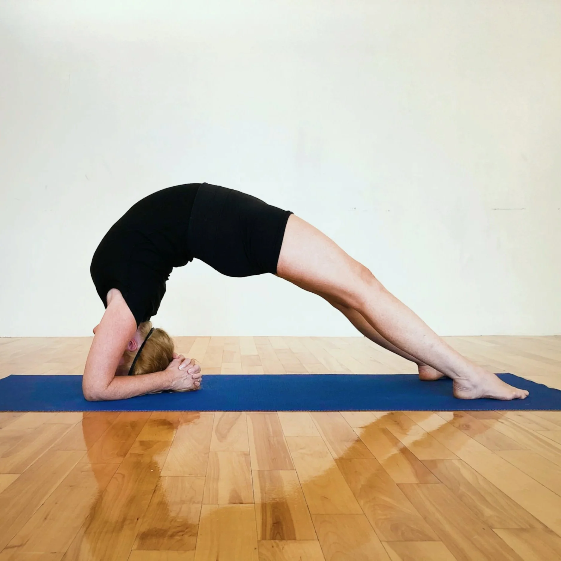 A person in black athletic clothing performing a yoga pose called downward dog on a blue mat in a studio with wooden flooring.