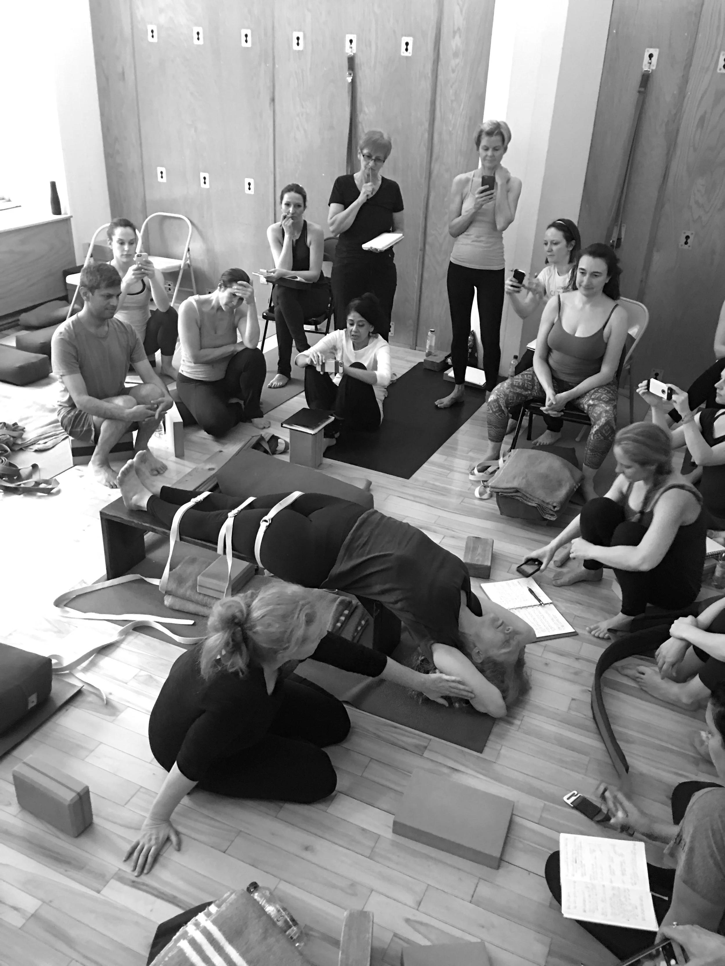 A group of people participating in a yoga or wellness class, with one person lying on a mat performing a pose while the instructor and others observe or record the session.