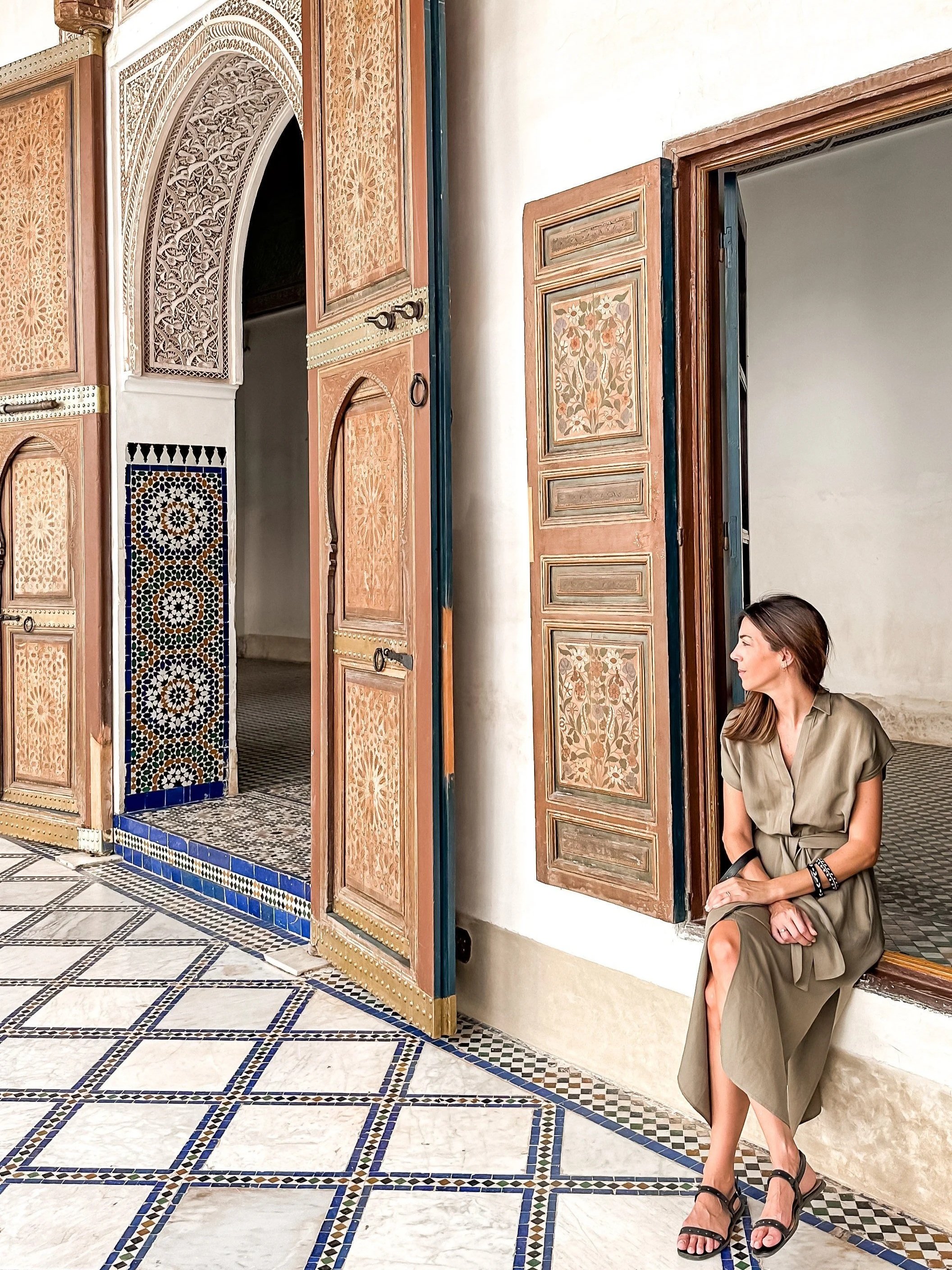 A woman in a beige dress sitting outside an ornately decorated building with colorful mosaic tiles and wooden panels.