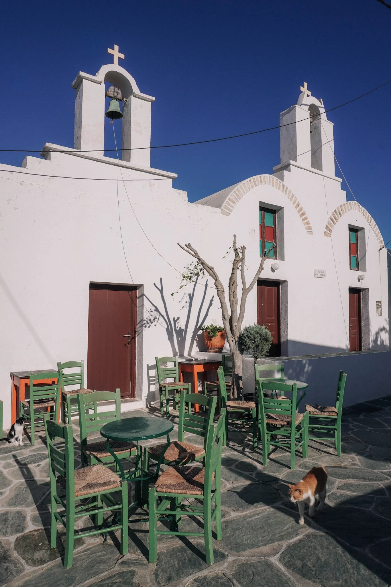 Little white church with brown doors and green tables and chairs in the center of Folegandros chora