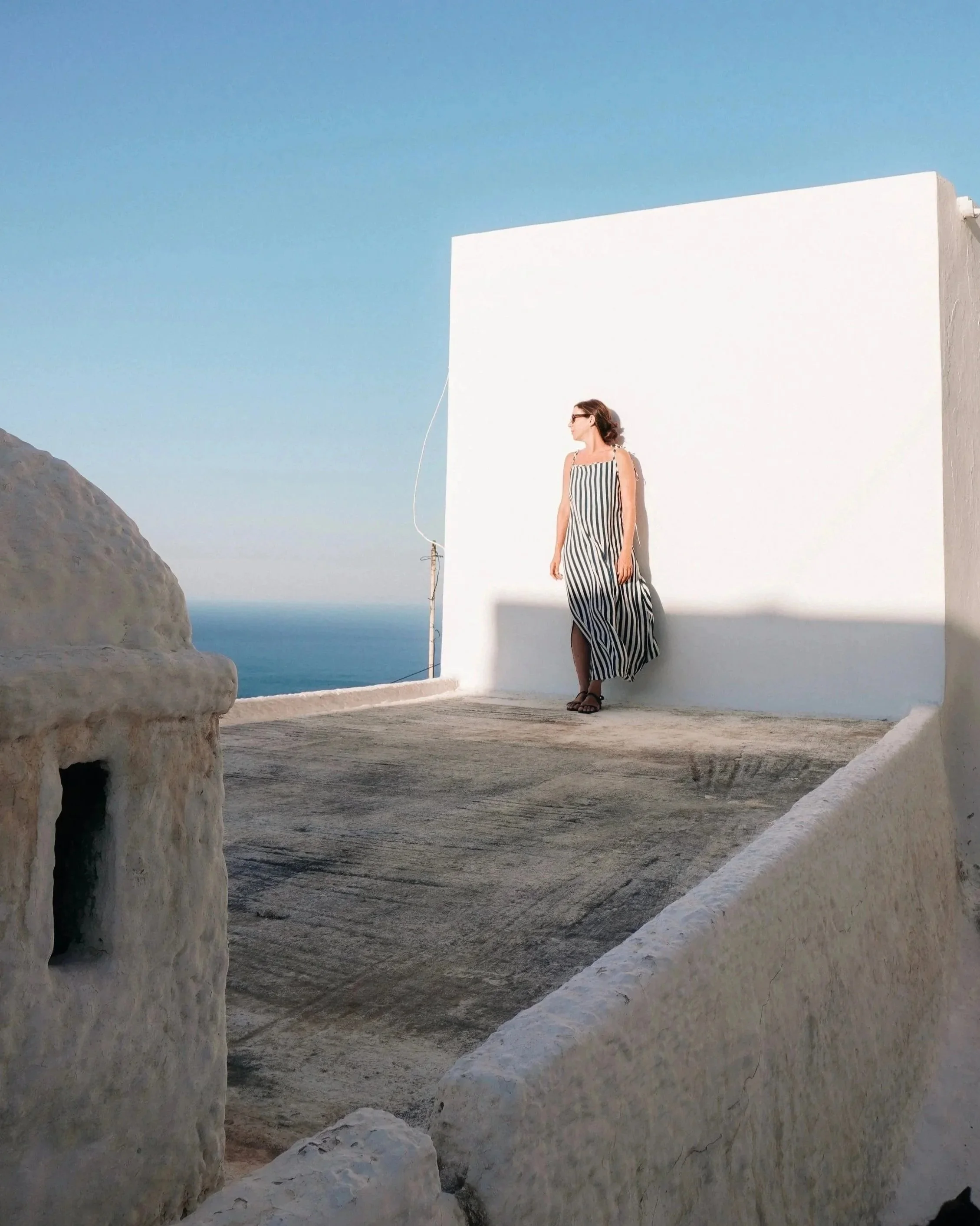 Woman in sunglasses and a striped dress standing against a white wall with a mountain and ocean in the background.
