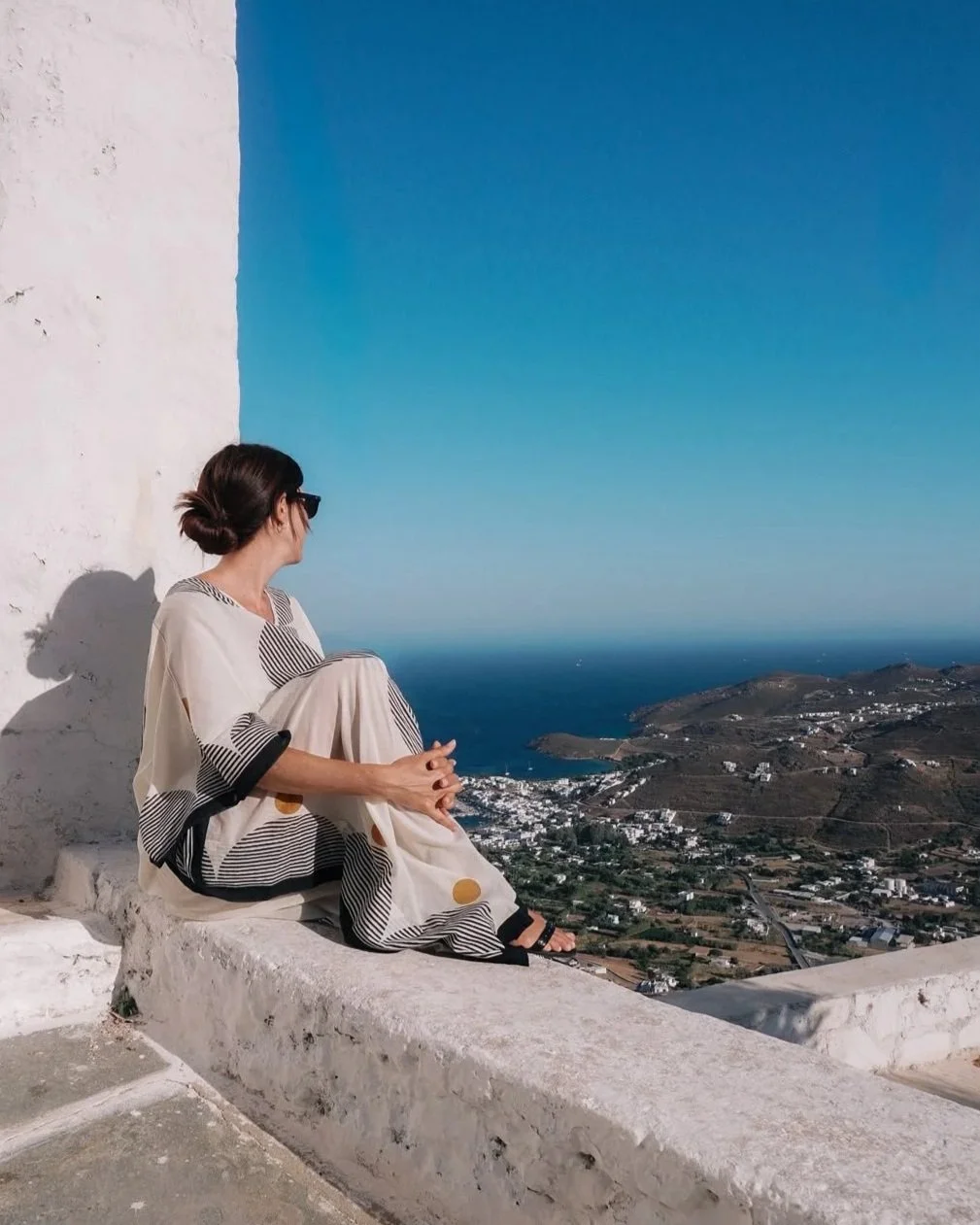 A woman sitting on a white wall overlooking a coastal landscape with blue sky and sea, wearing a striped dress and sunglasses, with her hair in a bun.