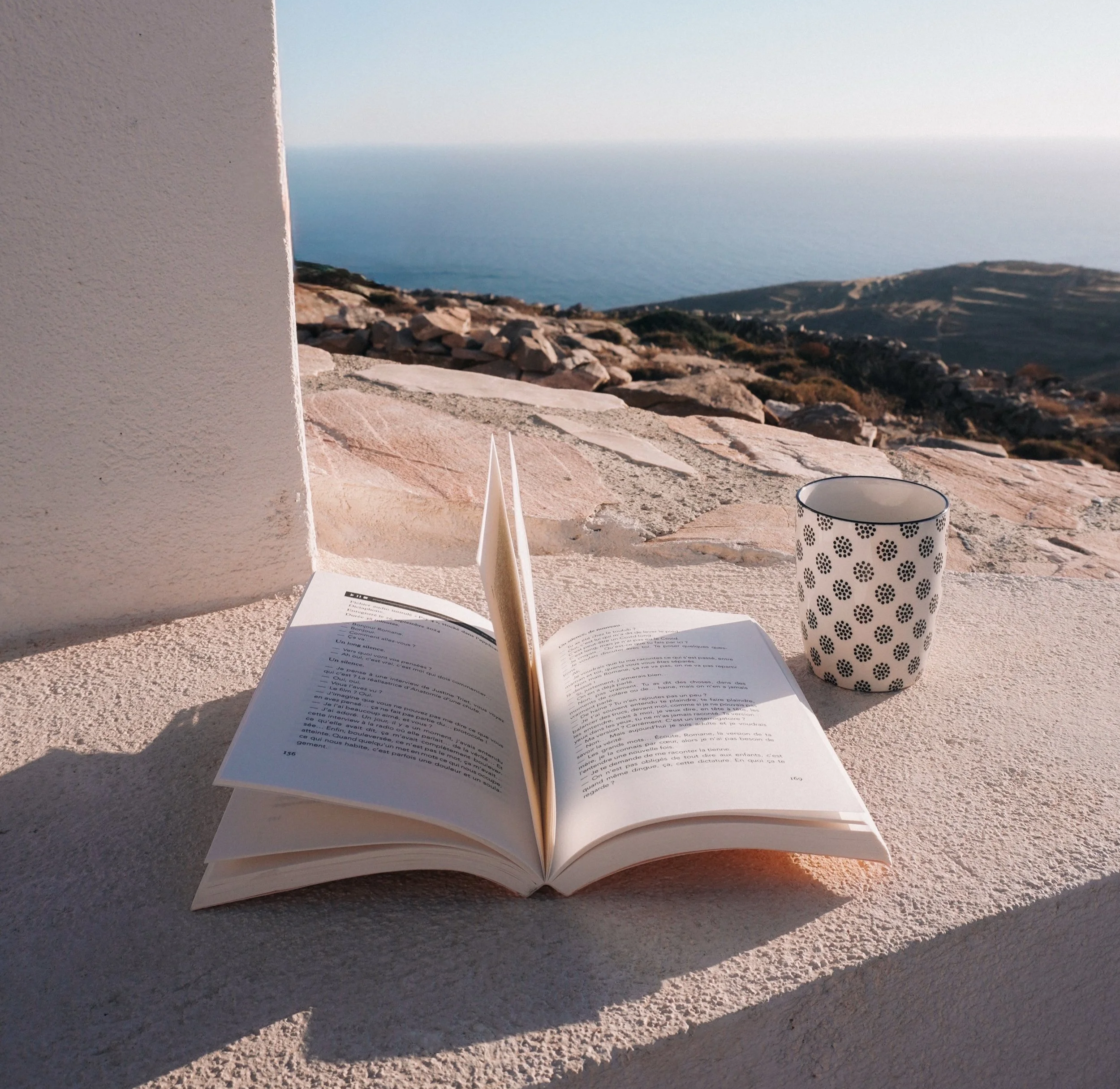 Open book with readable text, white and black polka dot mug, and white paper in sunlight on a concrete surface with a distant view of the ocean and rocky landscape.