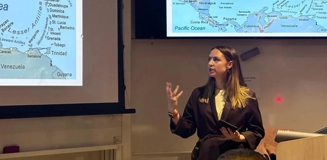 A woman giving a presentation about a world map focusing on the Caribbean and Central America, with a slide showing islands and countries including Venezuela, Trinidad, Barbados, and parts of Central America.