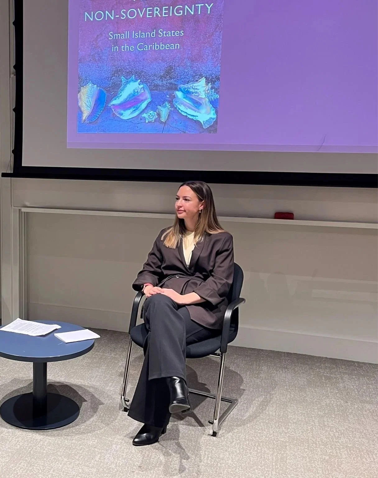 A woman in a dark suit sitting on a black chair in front of a presentation screen. The screen displays a slide titled 'Non-Sovereignty: Small Island States in the Caribbean' with a background image of seashells.