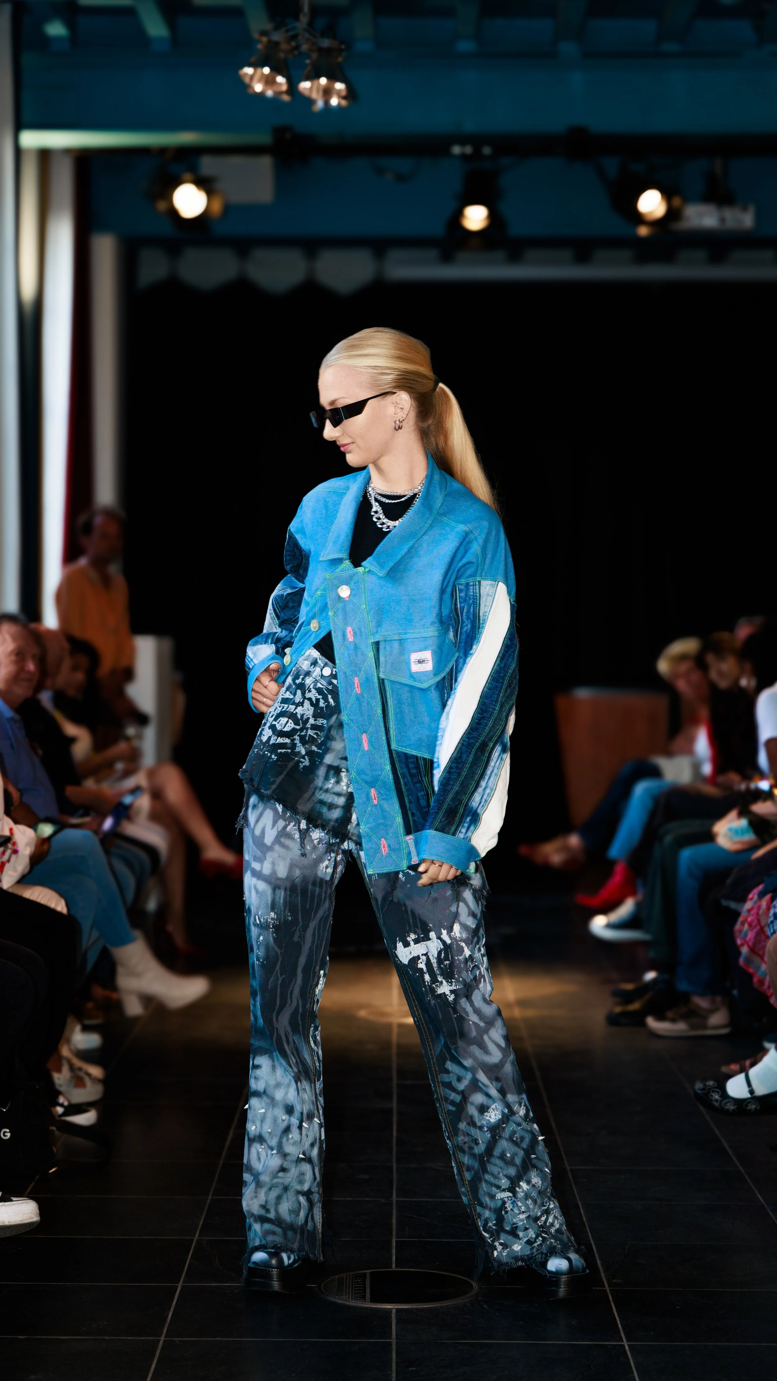 Model walking on runway in denim jacket, patterned pants, and sunglasses during fashion show.