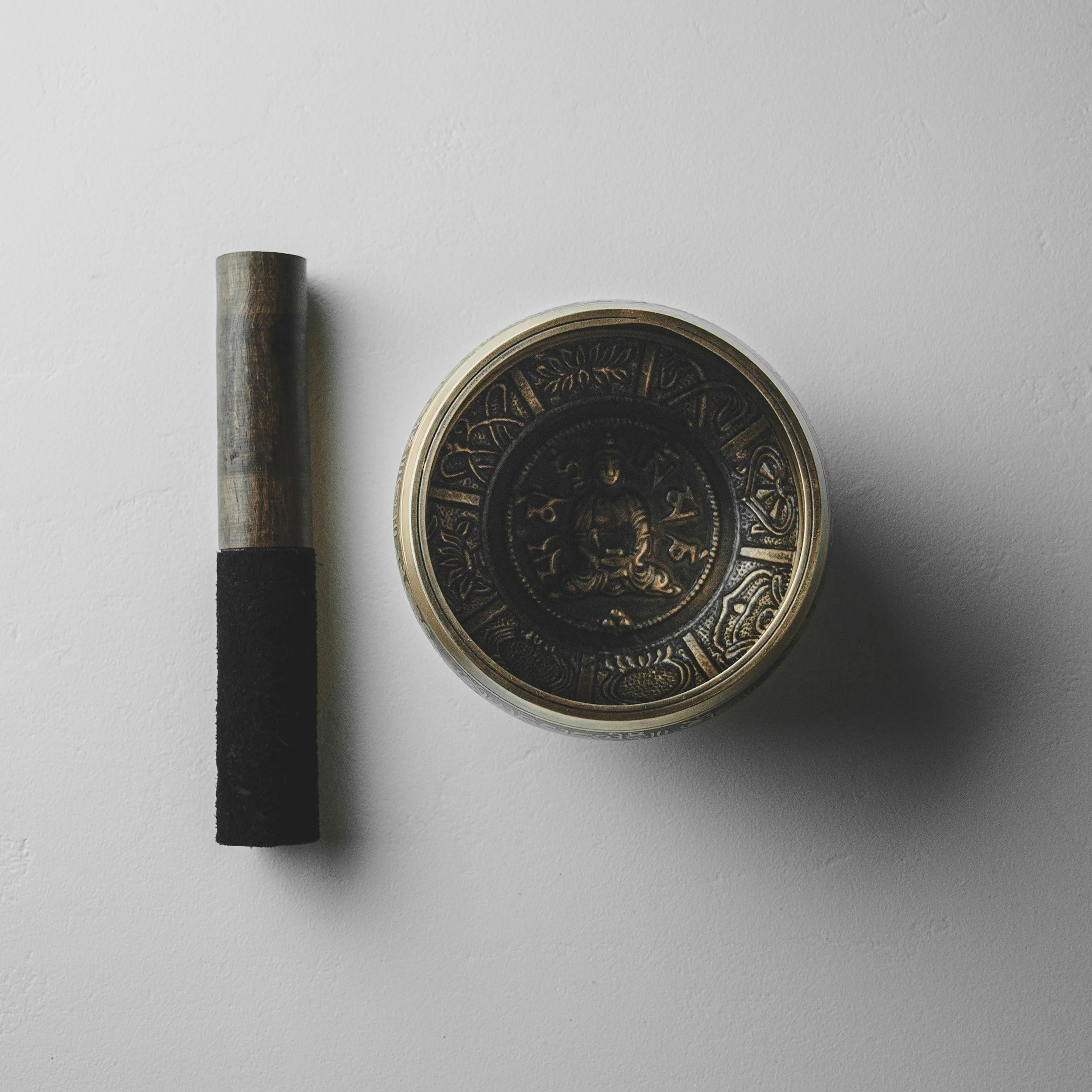 A lit incense stick with ash at the tip placed next to a round decorative container featuring Asian-inspired designs on a white surface.