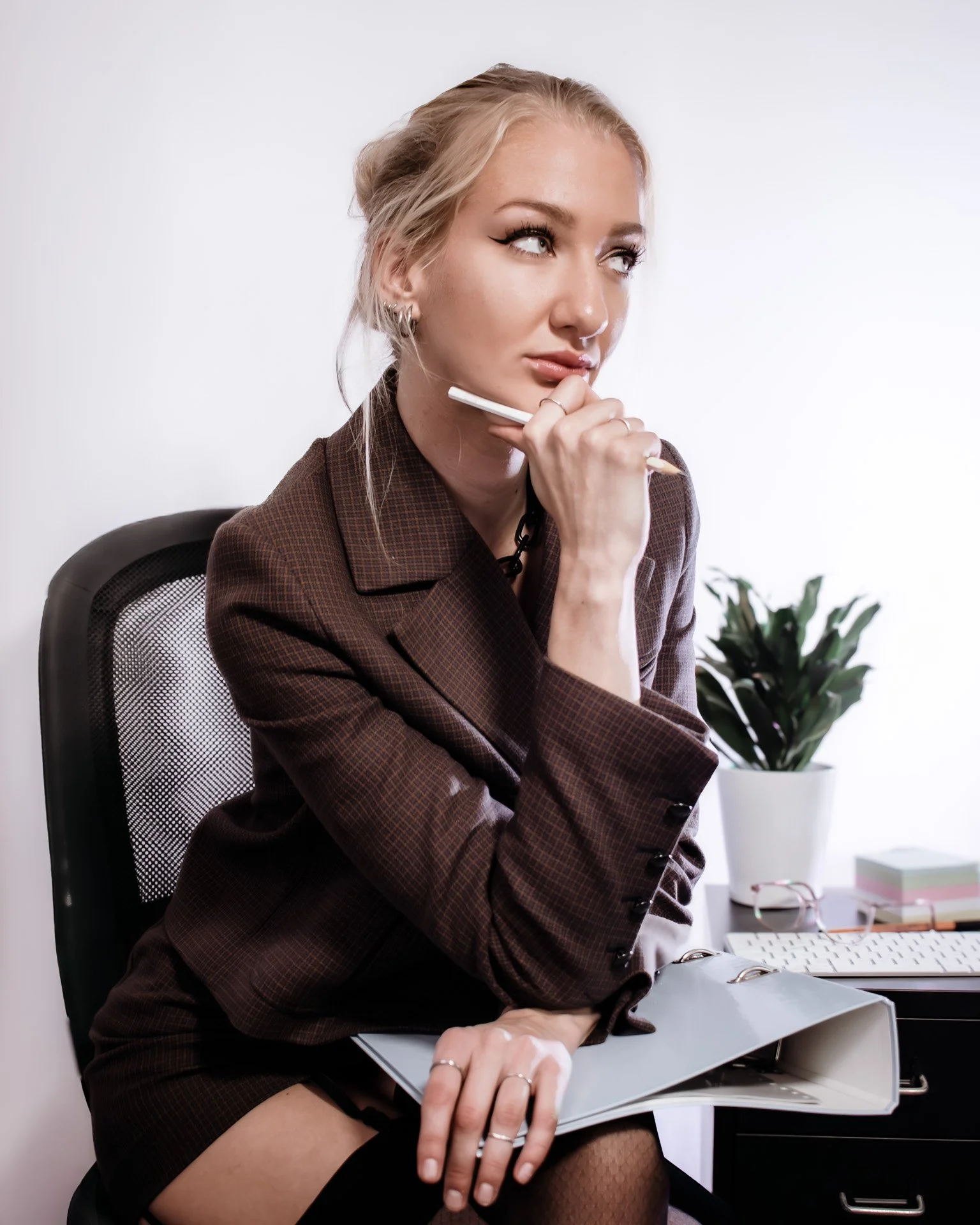 Woman with blonde hair and hoop earrings, dressed in a brown plaid blazer, sitting at a desk with a gel pen near her chin, holding an open folder, with a potted plant and office supplies in the background.