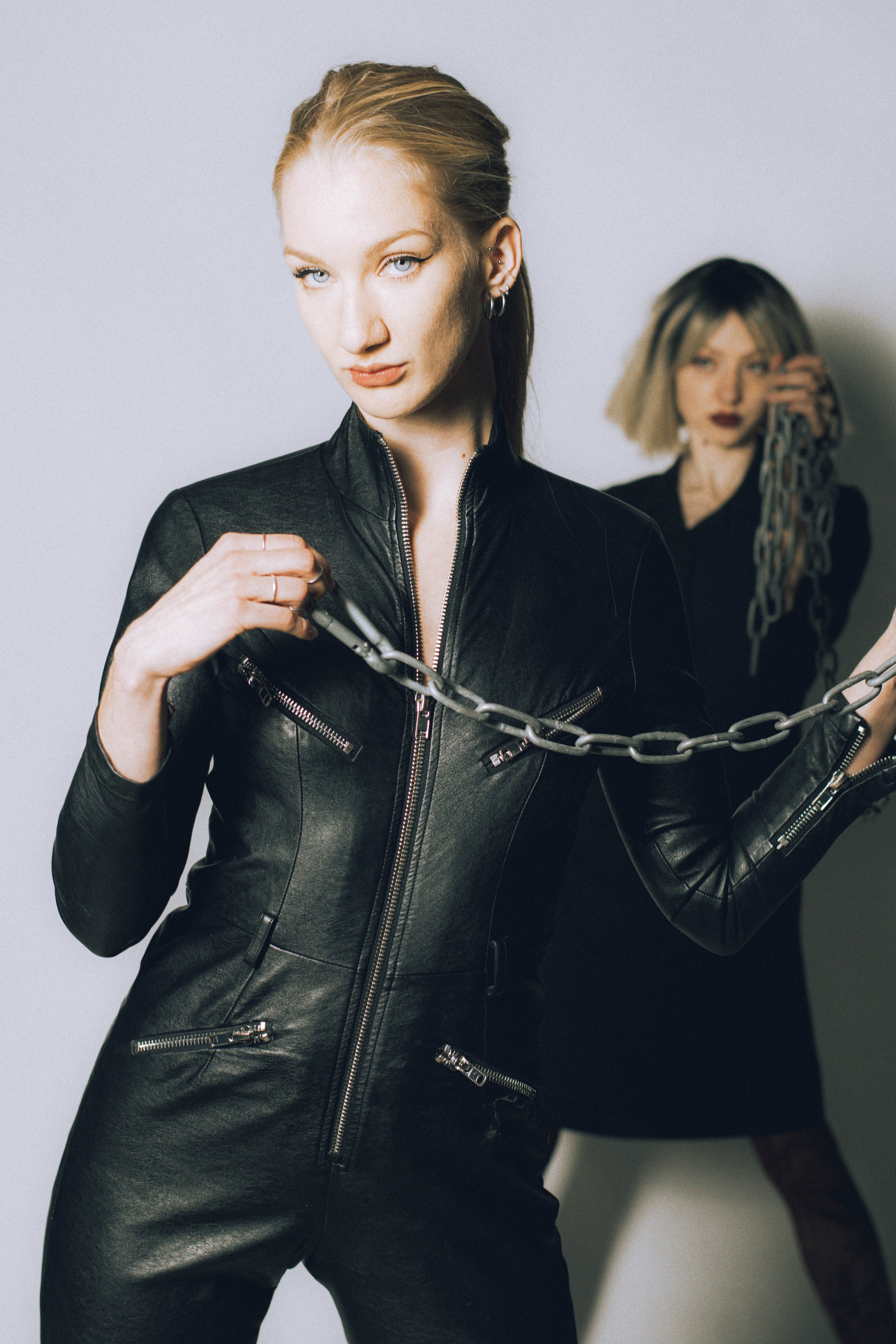Two women posing, with the woman in the front holding a chain. The woman in the front has blonde hair, wears a black leather jacket, and looks directly at the camera. The woman in the background has blonde hair with dark roots and is holding a chain near her face.