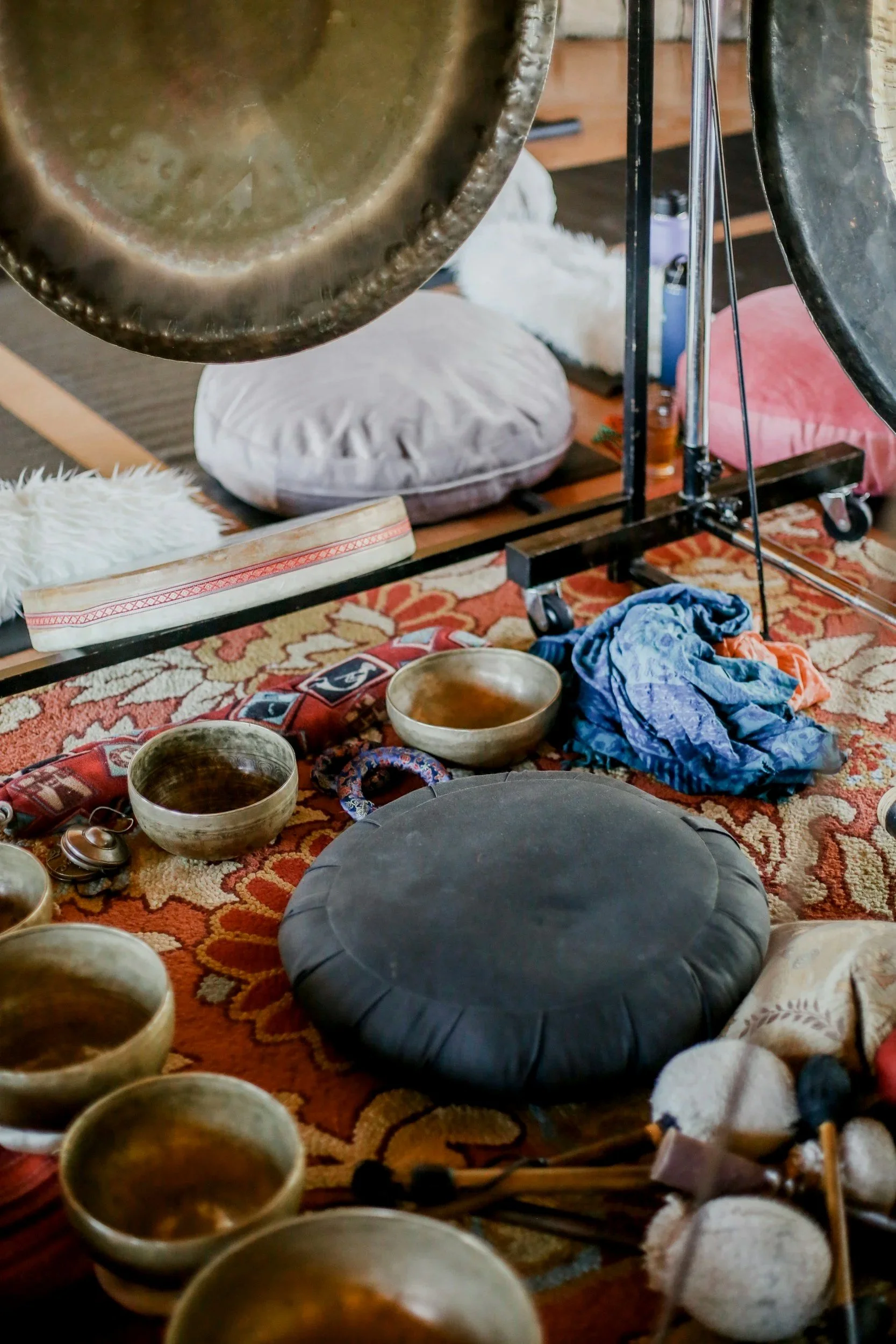 A collection of singing bowls, percussion mallets, a black meditation cushion, and scarves on a decorative area rug, with gongs and cushions in the background.