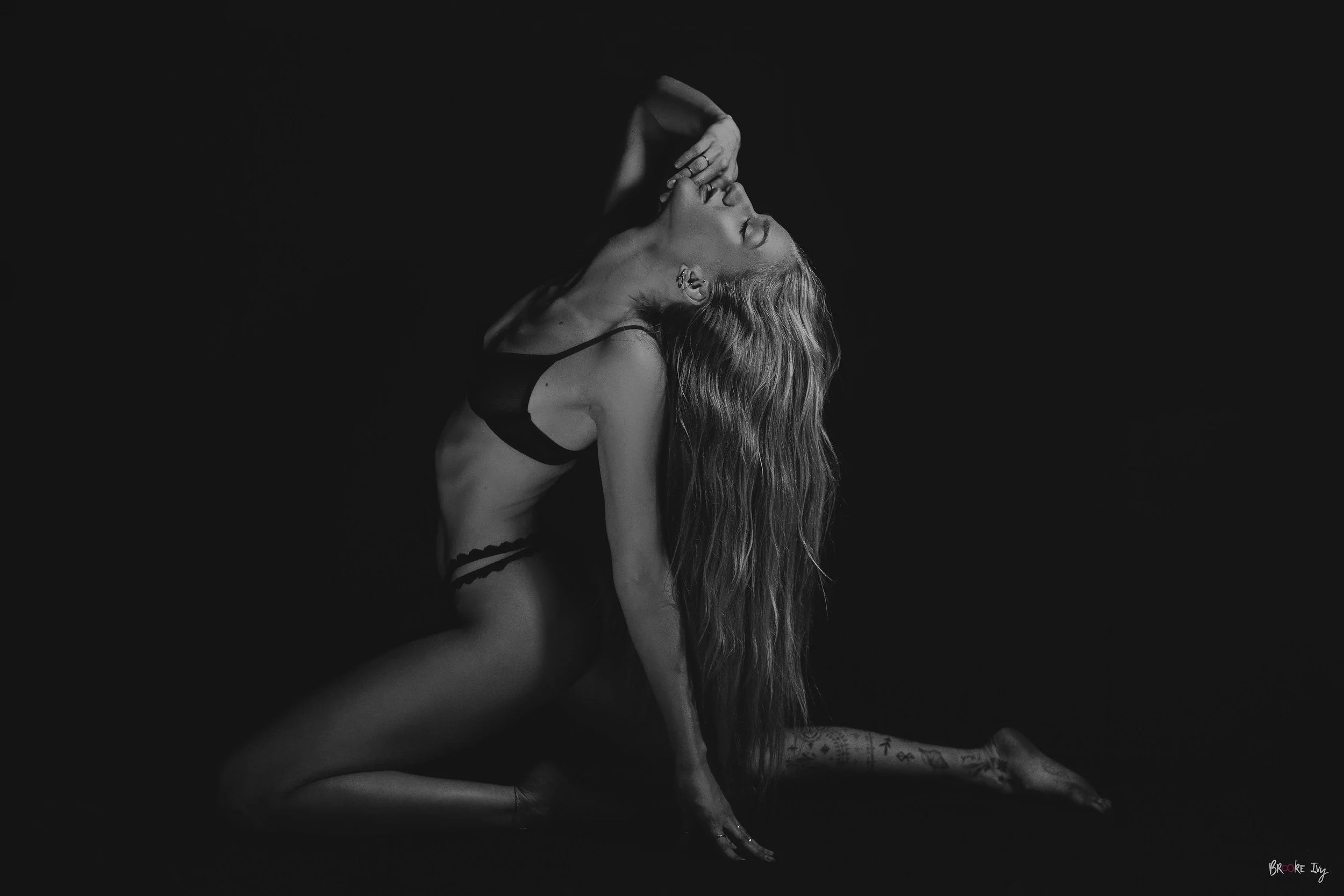 A woman with long wavy hair, wearing black lingerie, poses on her knees and toes against a black background, with one hand on the ground and the other touching her face in a dramatic, artistic pose.