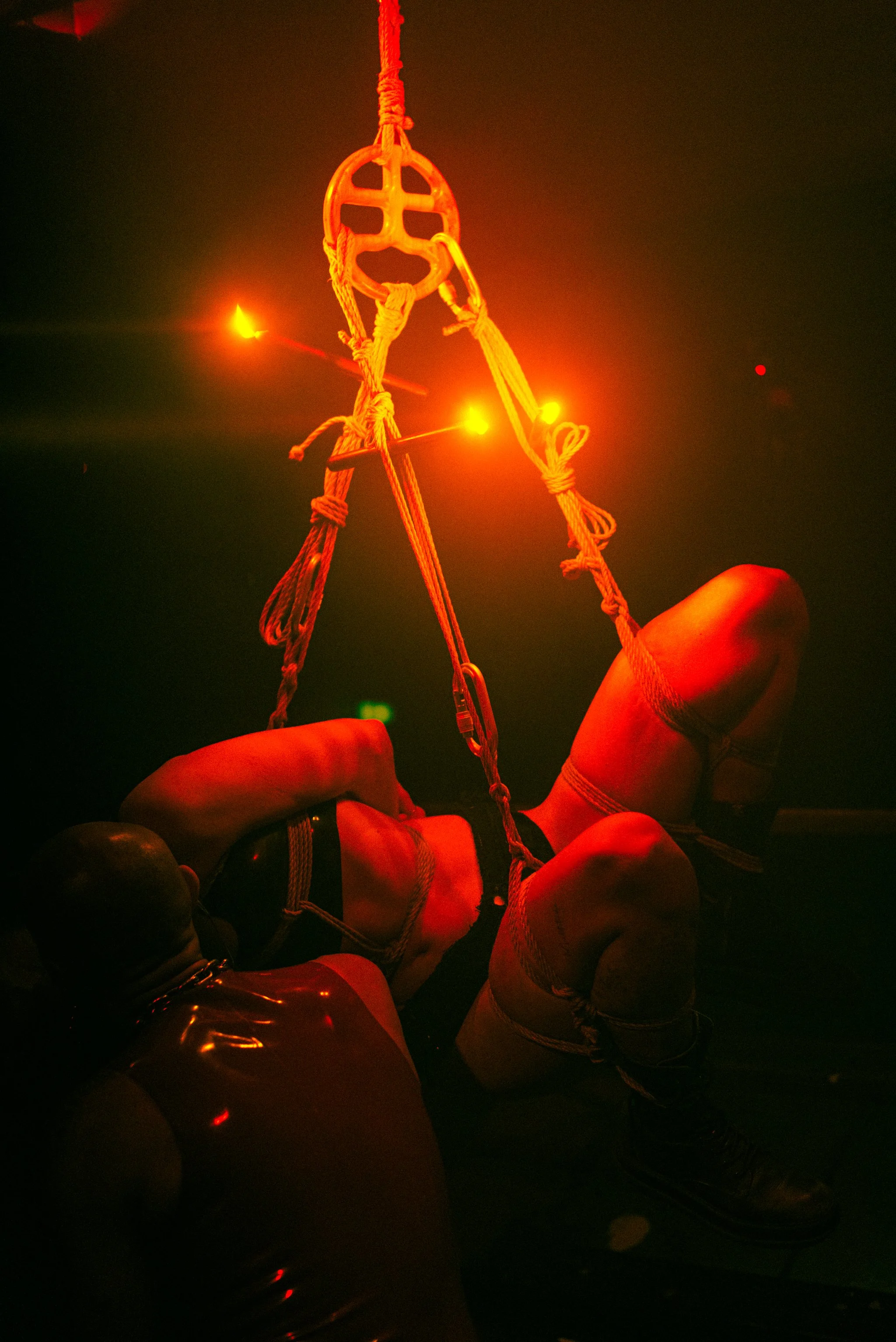 A person lying on the floor, wearing harnesses, with a rope and pulley system above them, illuminated by red lighting.