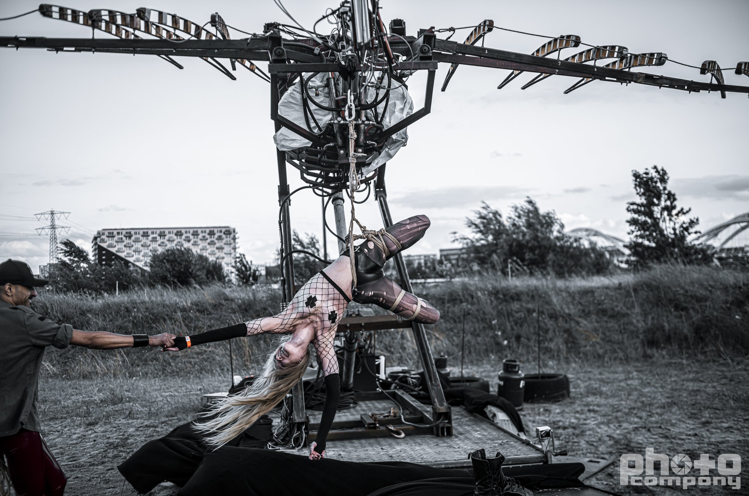 A woman with platinum blonde hair and fishnet clothing is hanging upside down from a large metal frame, being held by a man in a cap and dark clothing on an outdoor film set.