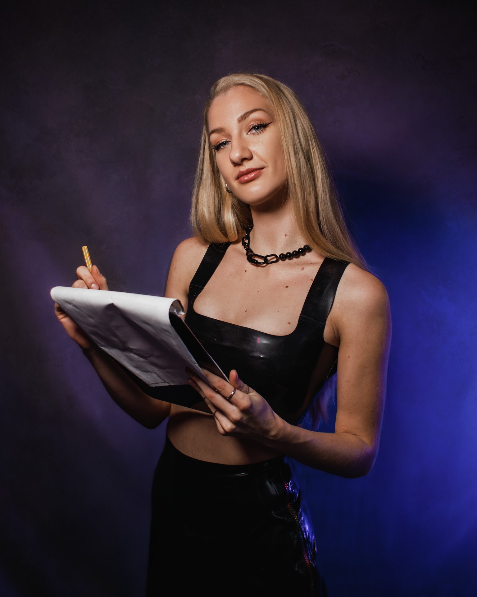 A young woman with long blonde hair, wearing a black sleeveless top and necklace, holding a notepad and pen, stands against a dark, purple-lit background.