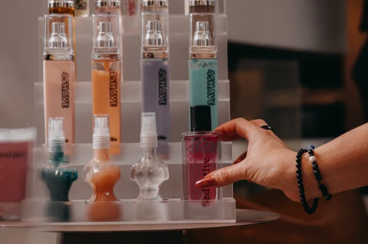 A hand reaching to pick up a pink lip gloss from a clear acrylic display with various lip glosses and nail polishes, some labeled '4SHAVE' and others with different colors.