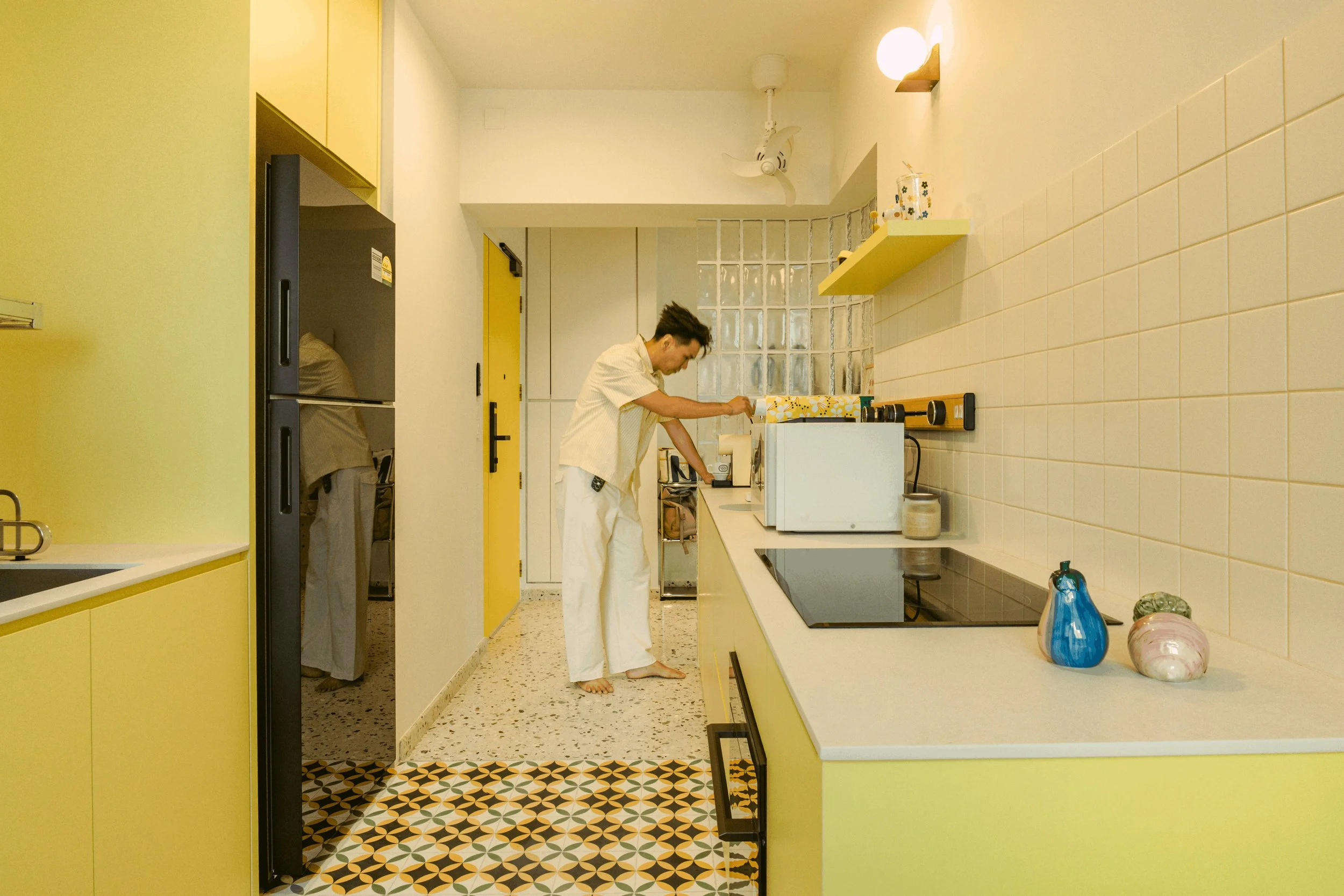 A person in a yellow and white checkered shirt and white pants standing in a kitchen pouring a drink from a cup. The kitchen has yellow cabinets, a white countertop, patterned tile floor, and decorative vases on the counter.