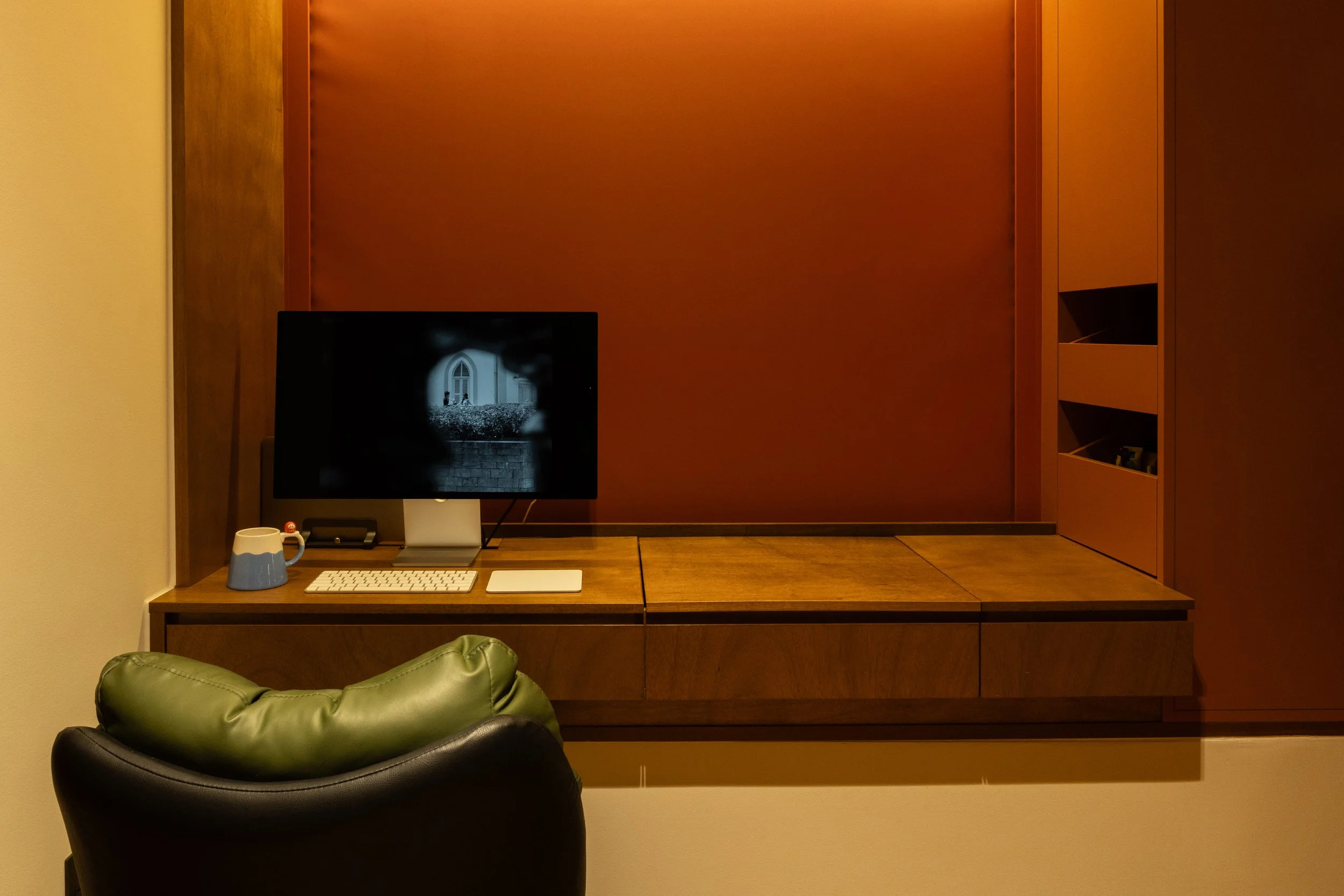 A computer workspace with a monitor showing a black and white image, a white keyboard, a mouse, and a mug on a wooden desk, with a green and black leather chair in front and a wooden wall with built-in shelving to the right.