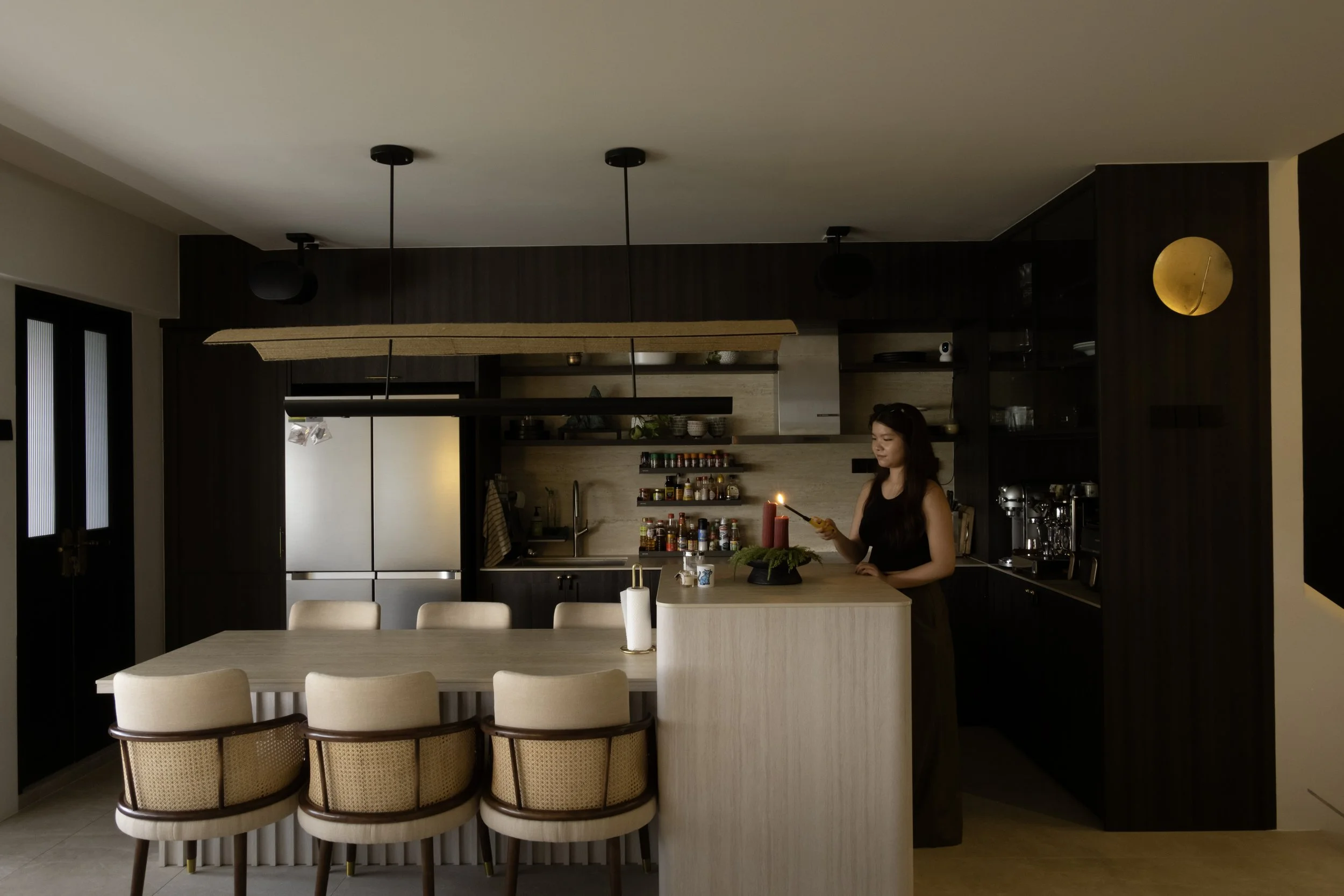 A woman in a black top preparing food in a modern, dark-paneled kitchen with pendant lights, a gold wall clock, and a dining table with beige chairs.