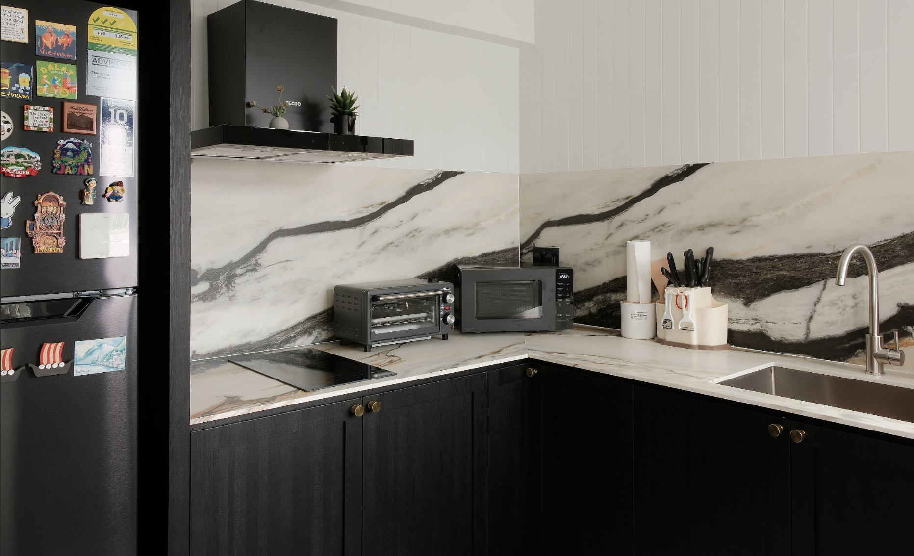 Modern kitchen with black cabinets, white and black marbled backsplash, stainless steel sink, small black oven and microwave, toaster oven, knife holder, paper towel holder, and a black refrigerator covered with colorful fridge magnets.