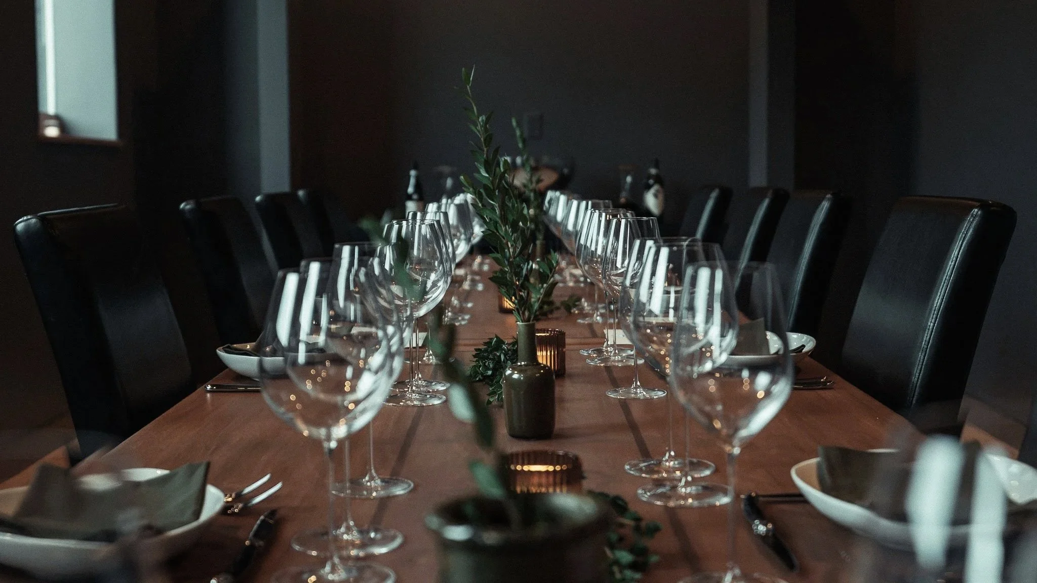 Interior of a modern, dimly lit restaurant with wooden floors and ceiling. Several tables and chairs are occupied by patrons, some watching TVs on the back wall. Outside the large windows, cars are parked.